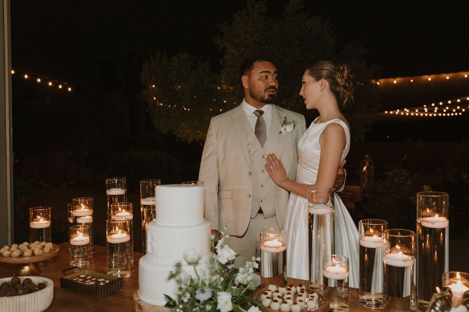 Couple shares a romantic moment at their candlelit wedding cake table surrounded by flickering candles and white florals.