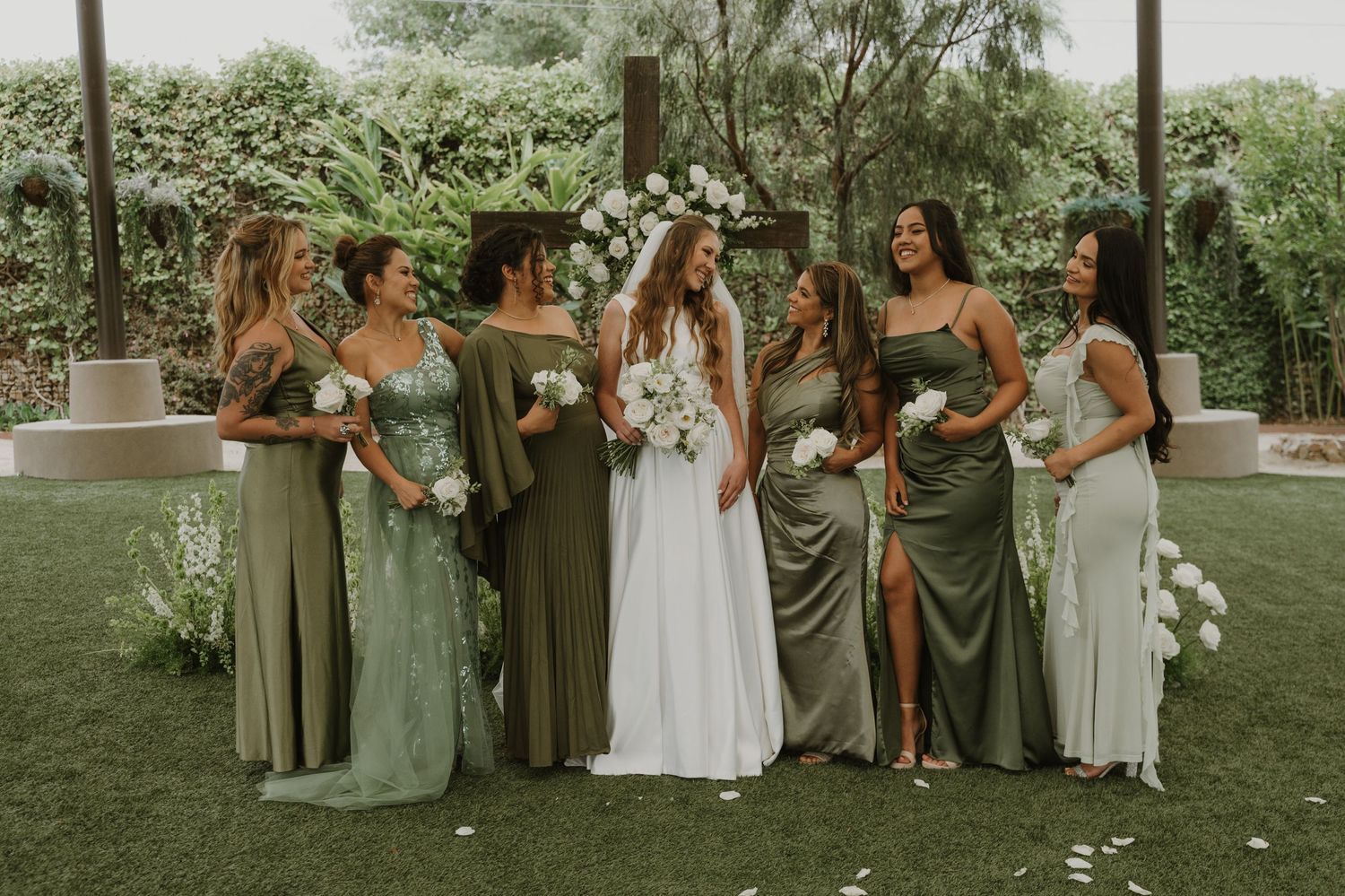 Bride and bridesmaids in sage green dresses posing outdoors at a garden wedding ceremony.