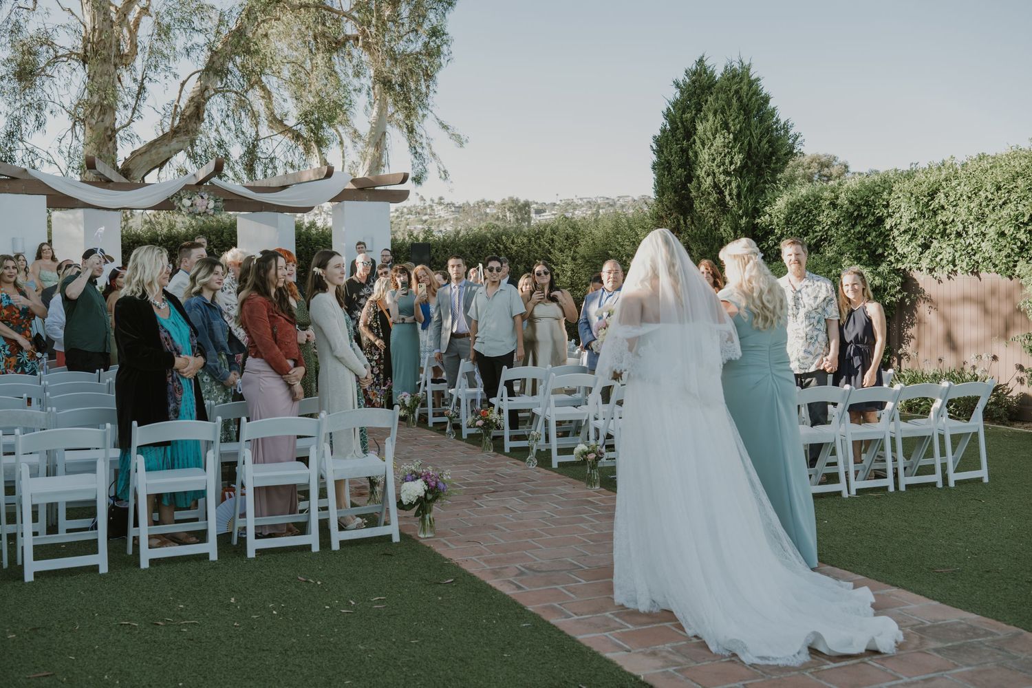 Bride in white gown and veil walks down aisle as guests stand and celebrate at outdoor garden wedding.