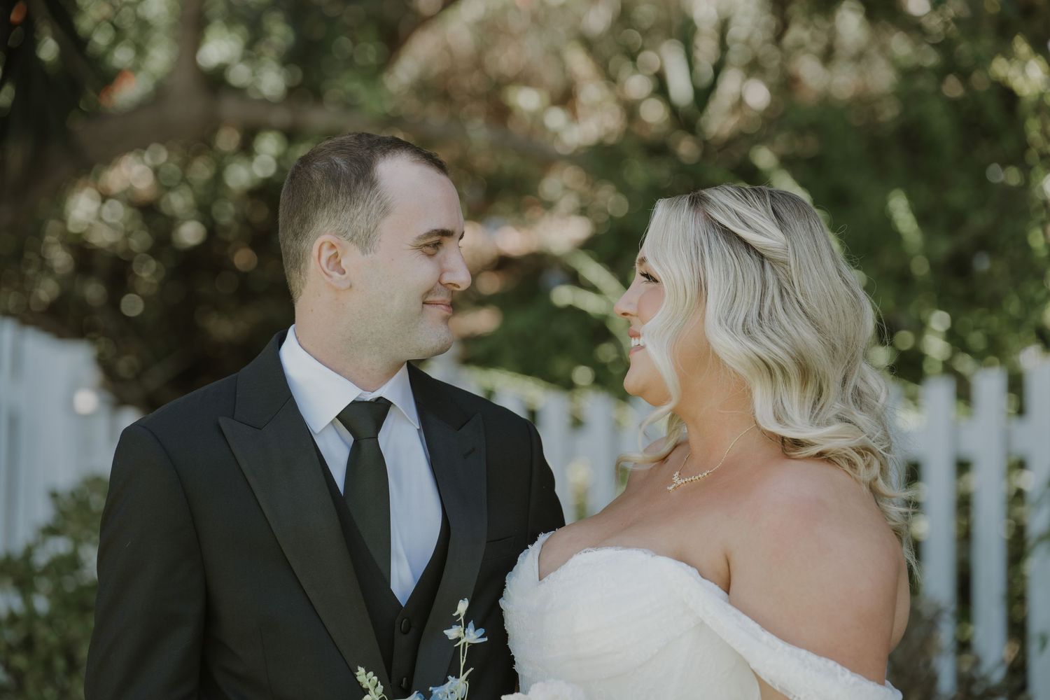 A bride and groom share a romantic glance outdoors, she in a strapless gown and he in a black suit with tie.
