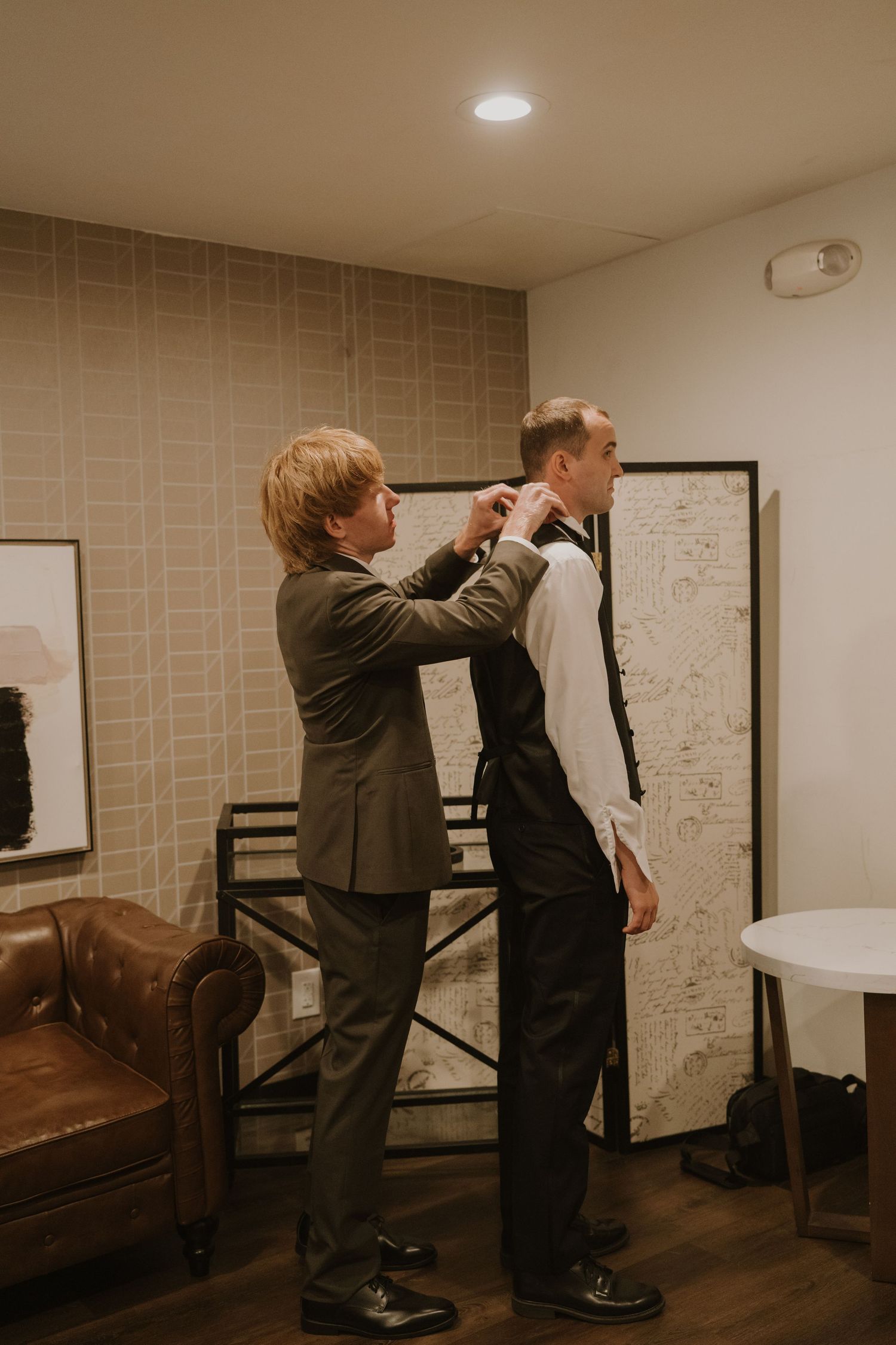 A groomsman helps adjust the groom's tie and collar while preparing for a wedding in an elegant room.