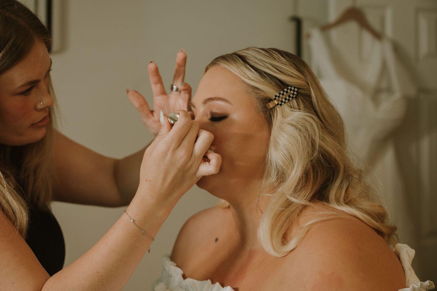A makeup artist applies finishing touches to a blonde bride wearing a hair accessory on her wedding day.