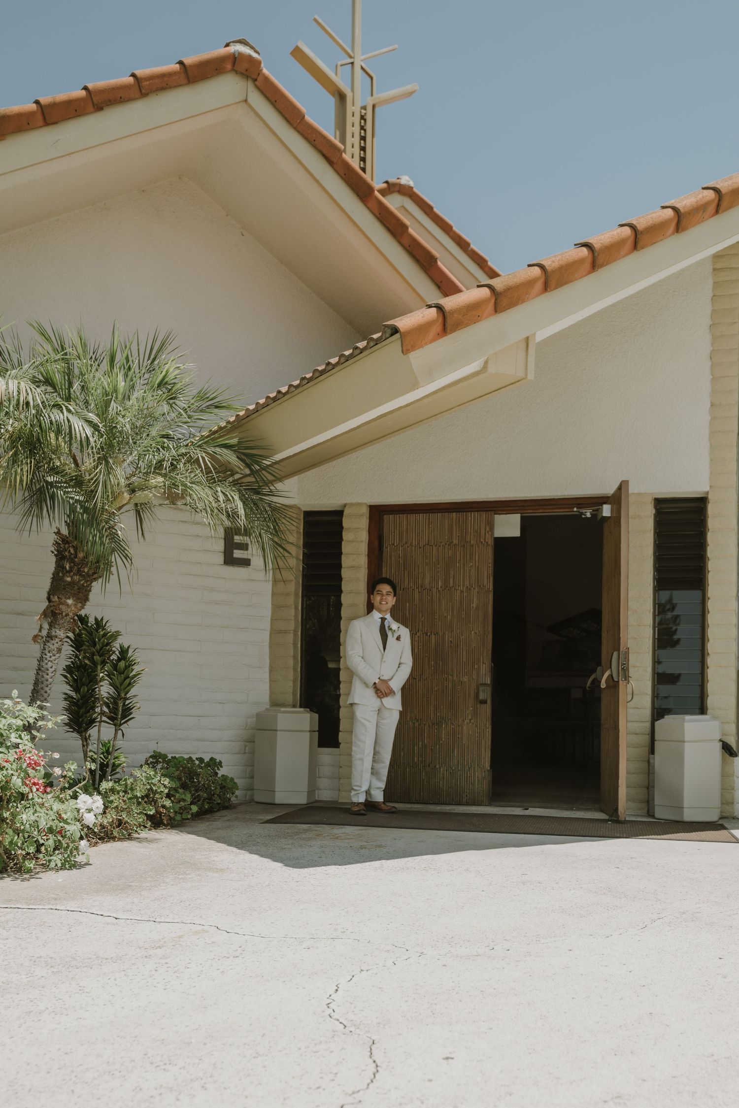 Groom in white suit standing at church entrance with palm trees and terracotta roof on sunny wedding day.