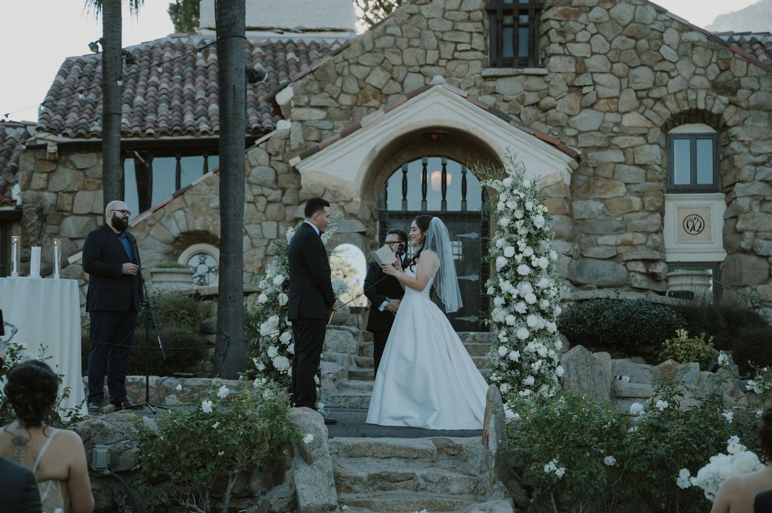 An outdoor wedding ceremony at a rustic stone chapel adorned with white floral arrangements.