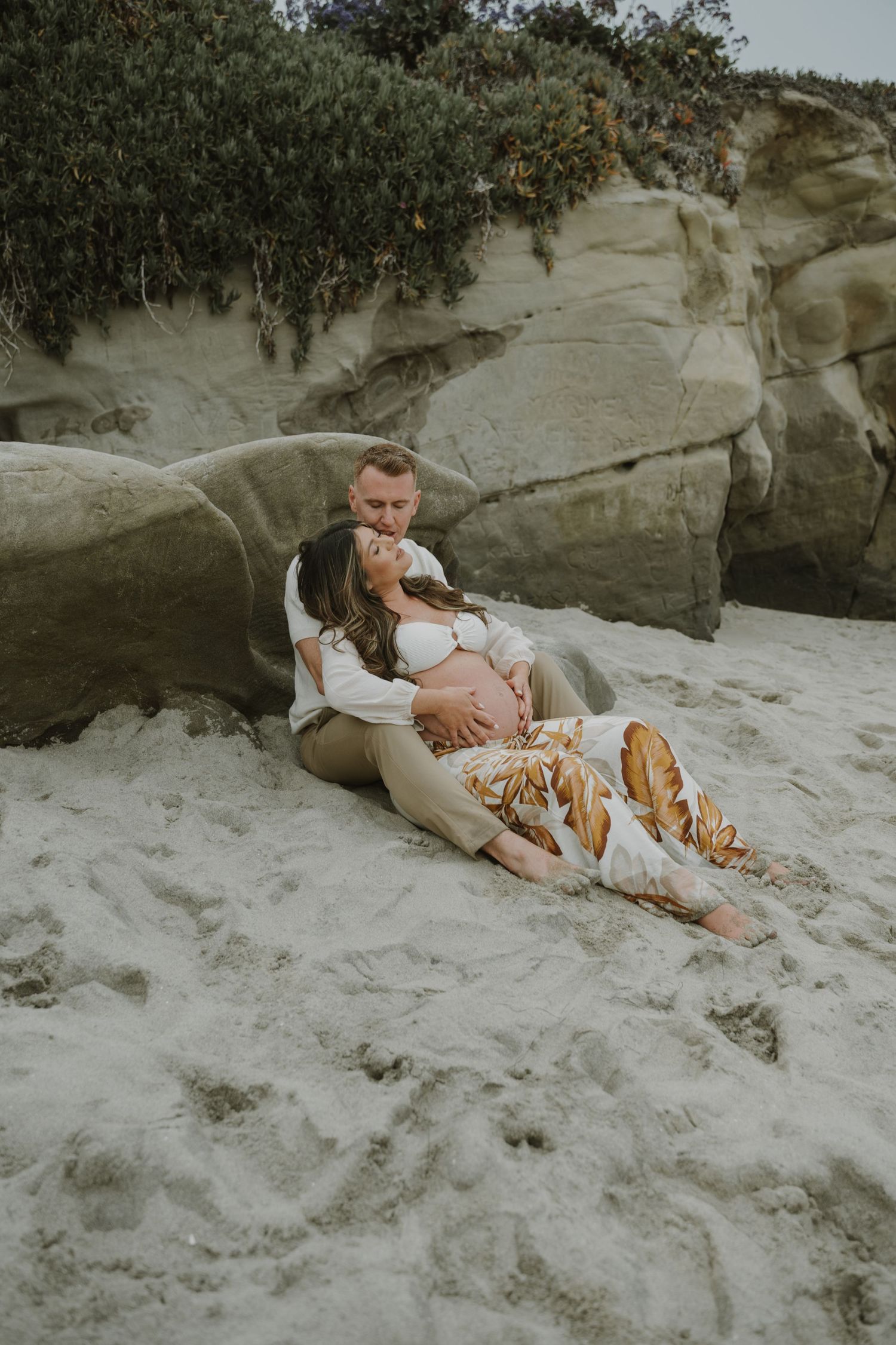 A maternity photoshoot on a sandy beach with white flowing dresses and natural rock formations.