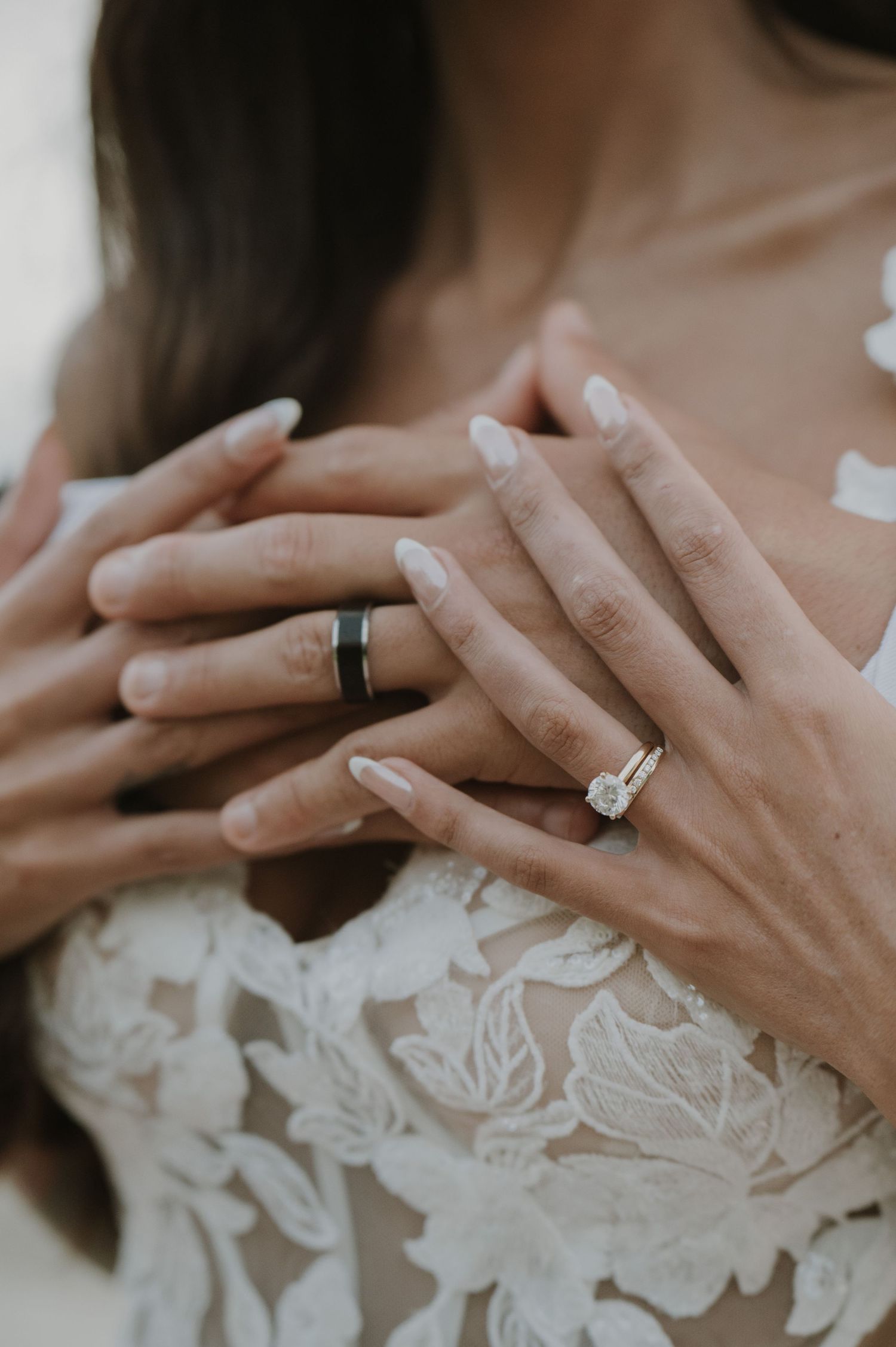 Detail shot of hands displaying wedding rings against white lace dress fabric.