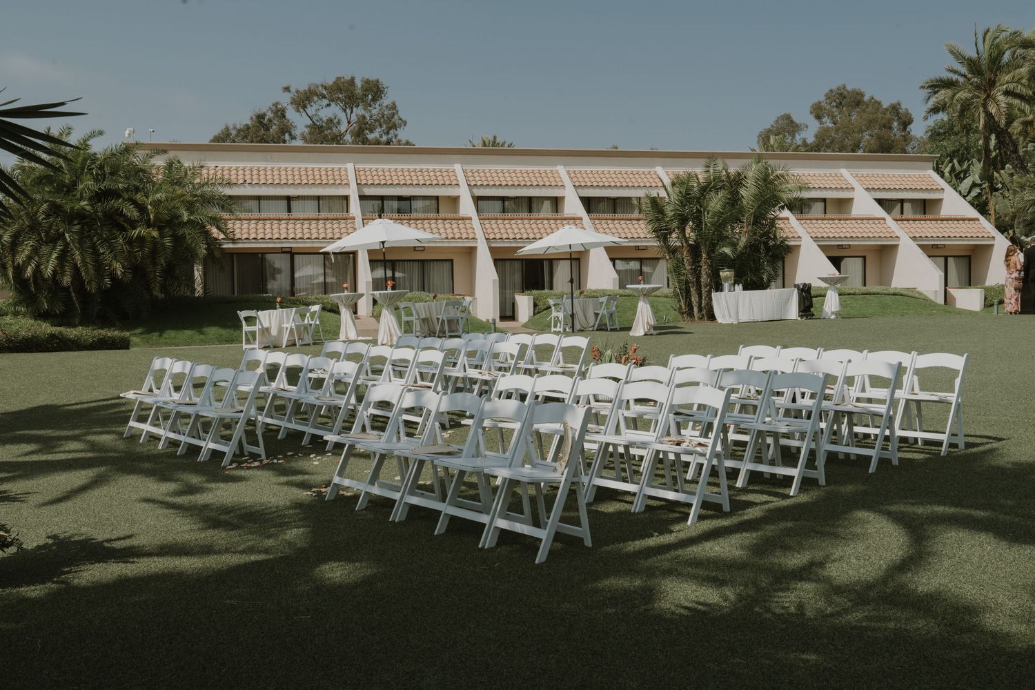 White ceremony chairs arranged in rows on a lawn with palm trees in the background.
