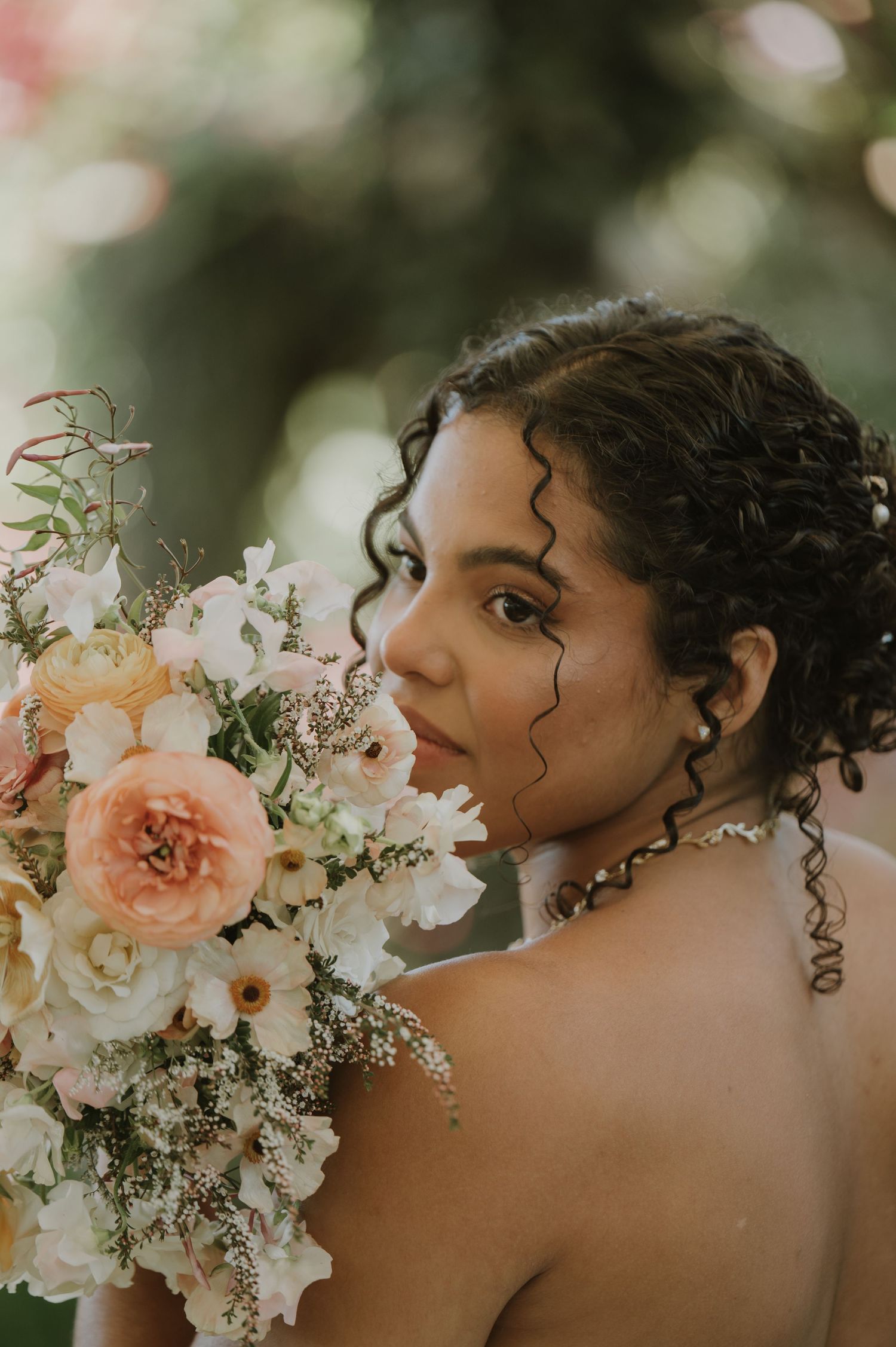 Bride with curly hair holds a peach and white floral bouquet, looking over her shoulder in a elegant outdoor portrait.