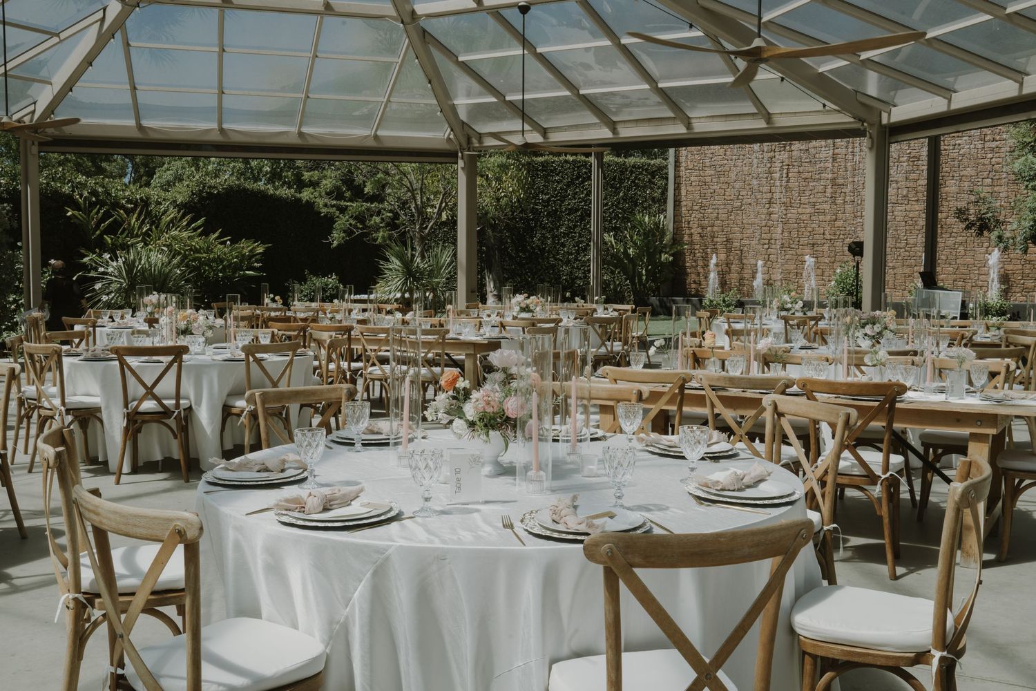 Elegant wedding reception in a glass greenhouse with round tables, white linens, and wooden cross-back chairs.