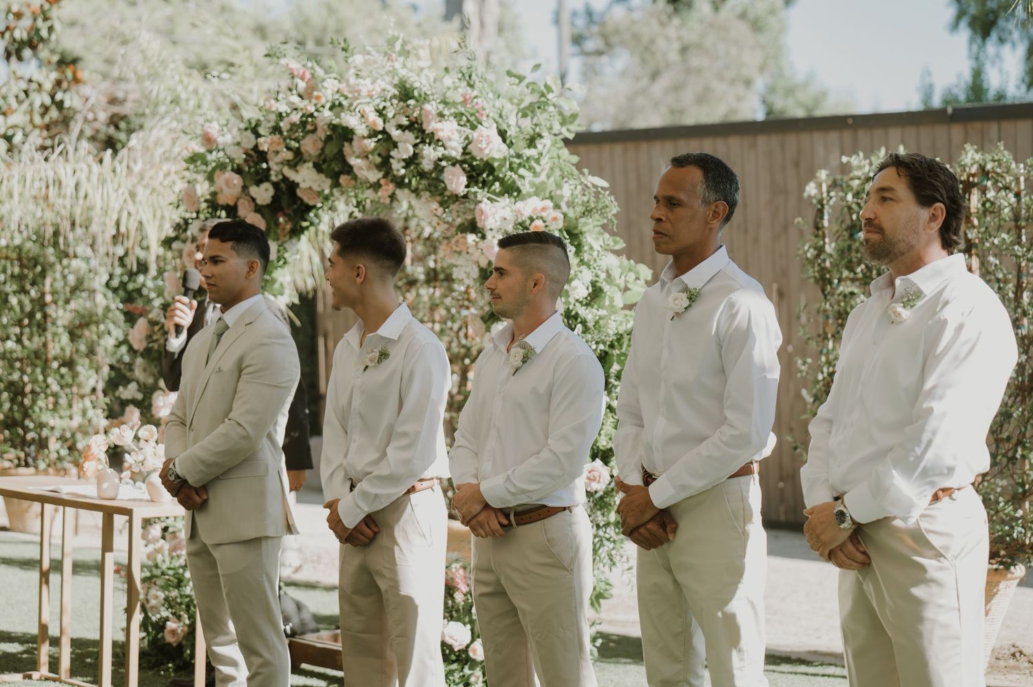 Four groomsmen in white shirts and khaki pants stand at outdoor wedding ceremony with floral arch backdrop.