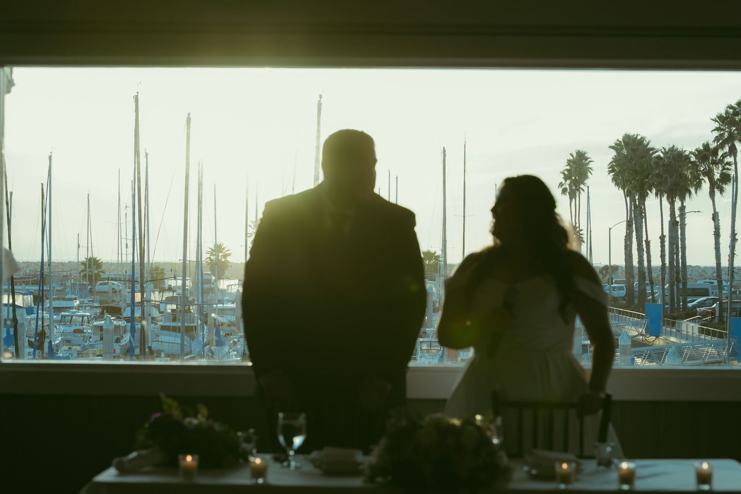 Couple silhouetted against sunset at waterfront restaurant with marina and palm trees in background.