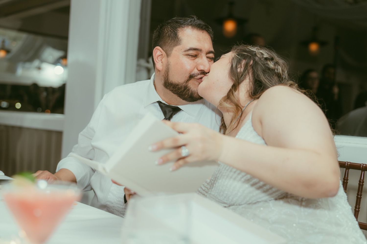 Bride and groom share a kiss while opening wedding gifts at their reception dinner table.