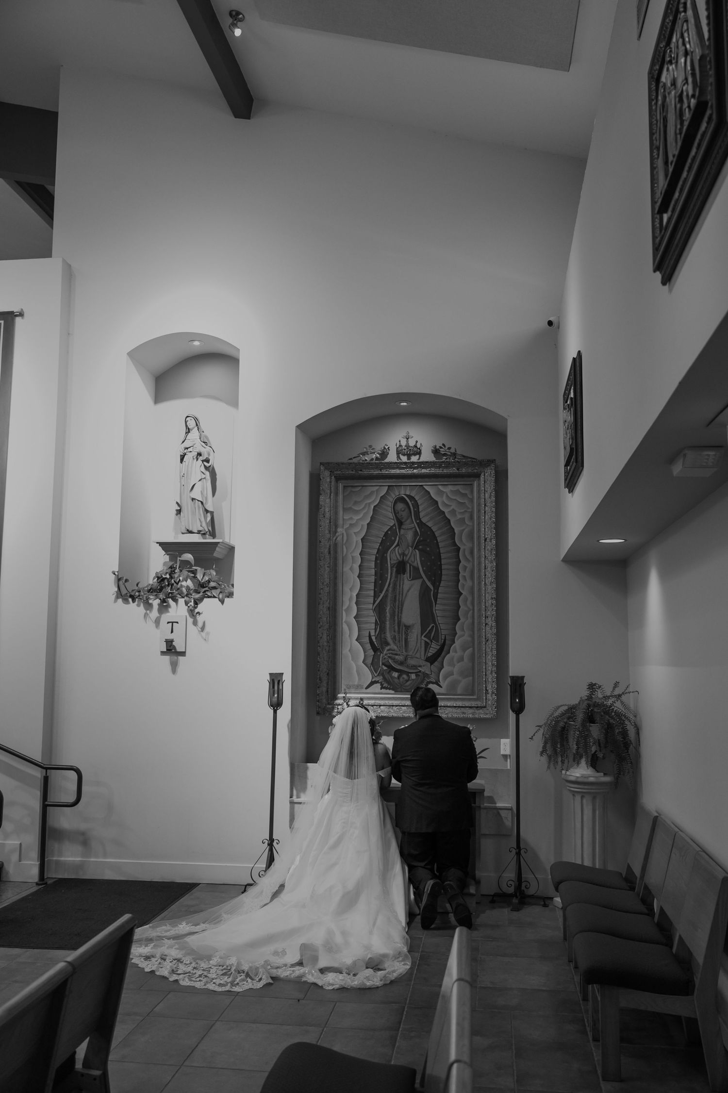 Bride and groom kneeling in prayer before Virgin of Guadalupe altar.
