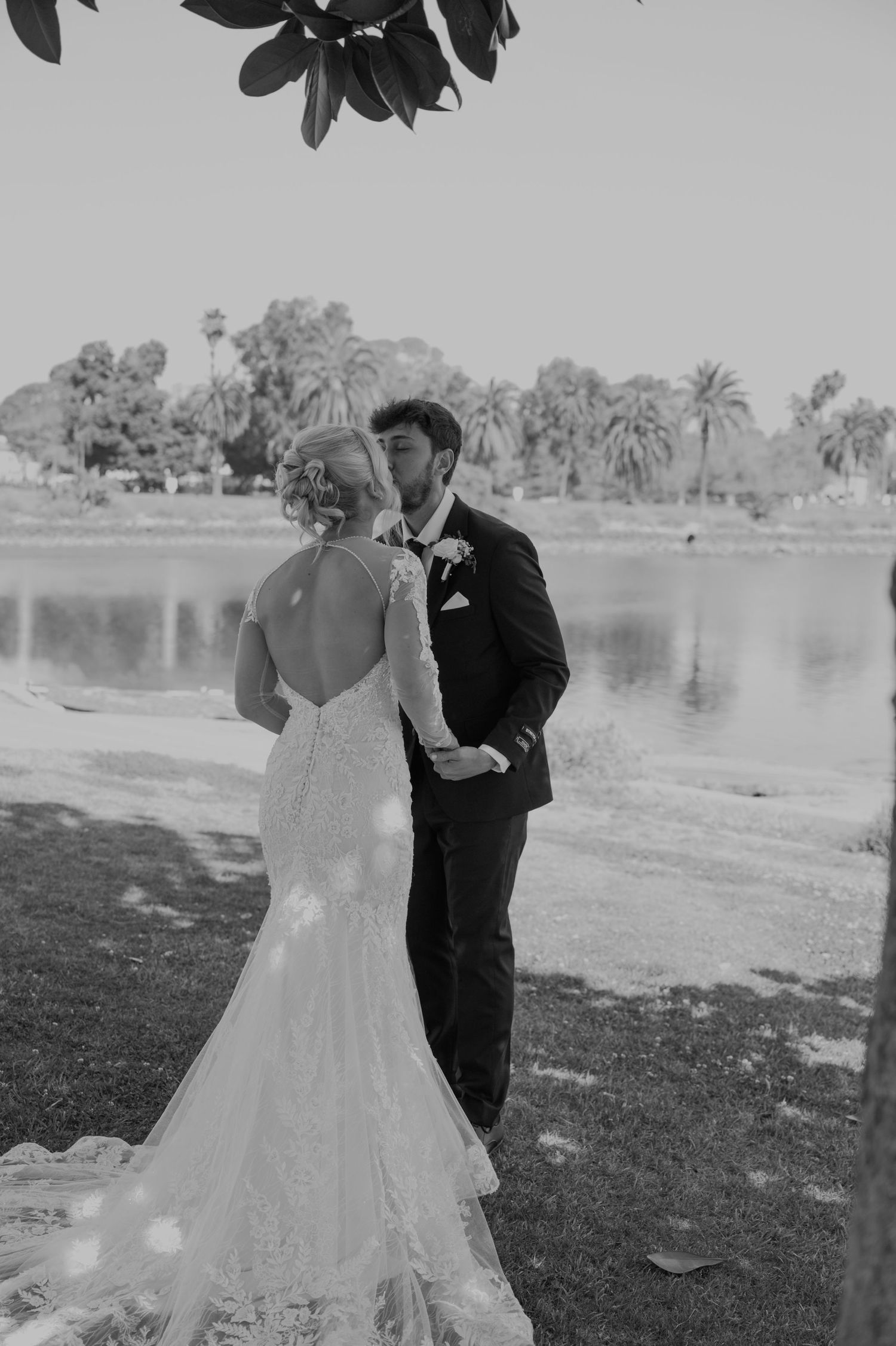 Black and white photo of a bride and groom sharing a kiss by a serene lake, surrounded by palm trees on their wedding day.