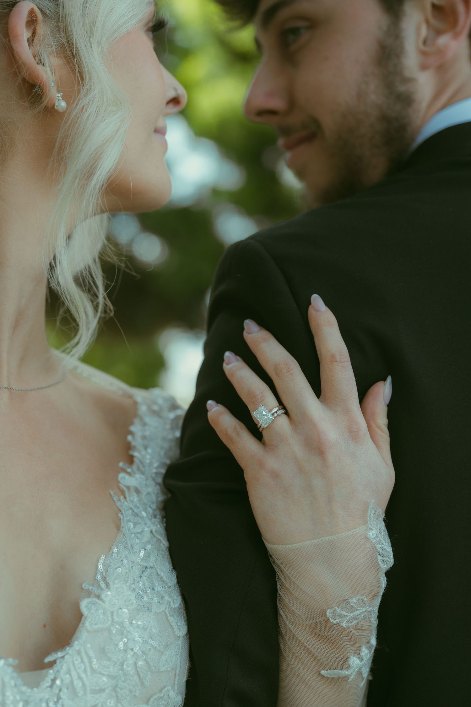 Bride in lace wedding dress rests hand on groom's suit, showcasing diamond wedding ring set.