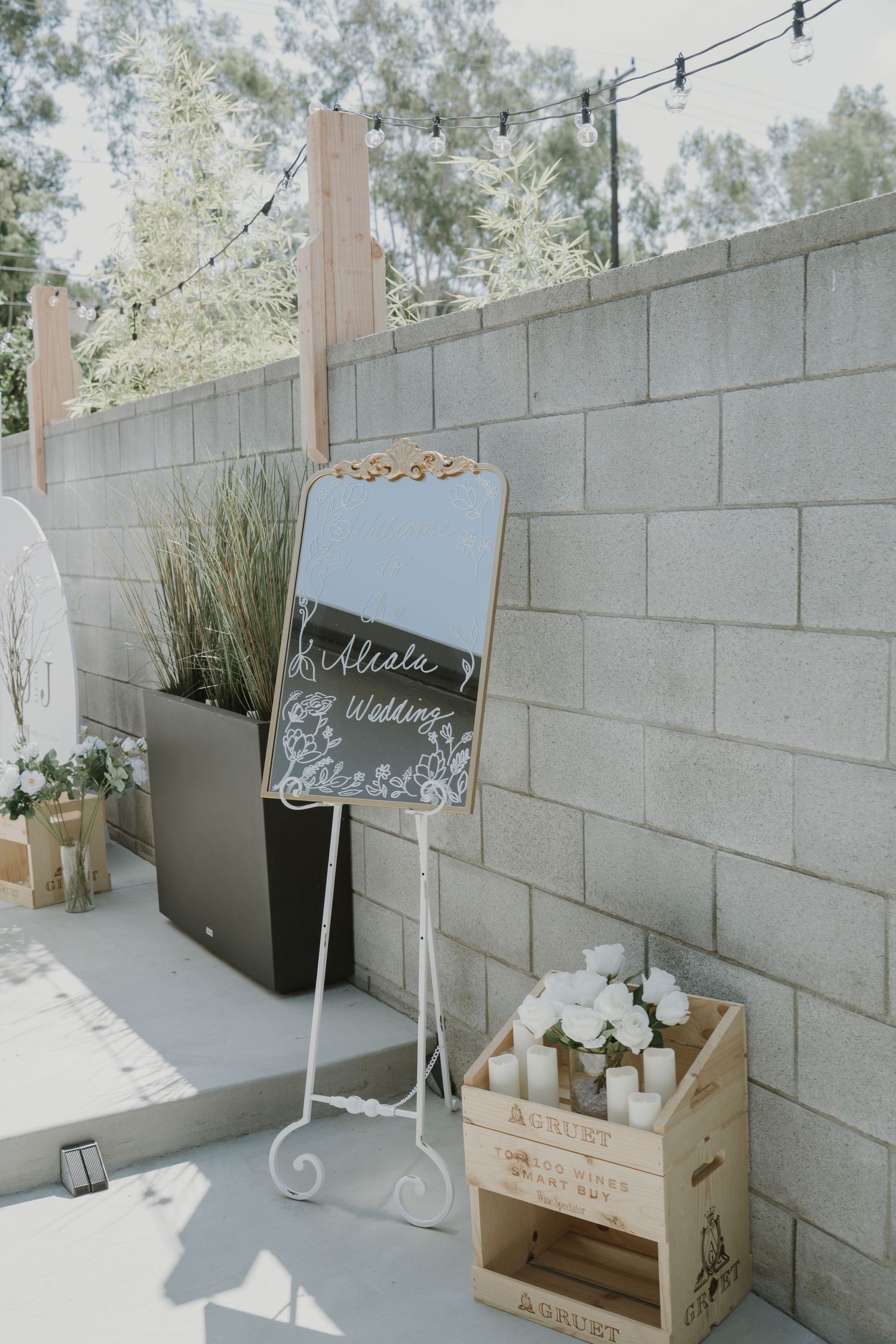 Elegant wedding welcome mirror sign on white easel with white roses in wooden crate against stone wall.