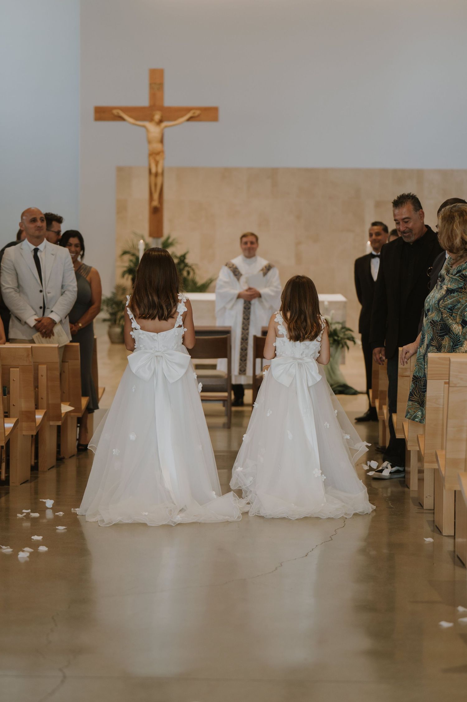 Two flower girls in white dresses with bows walk down the aisle at a Catholic church wedding ceremony.