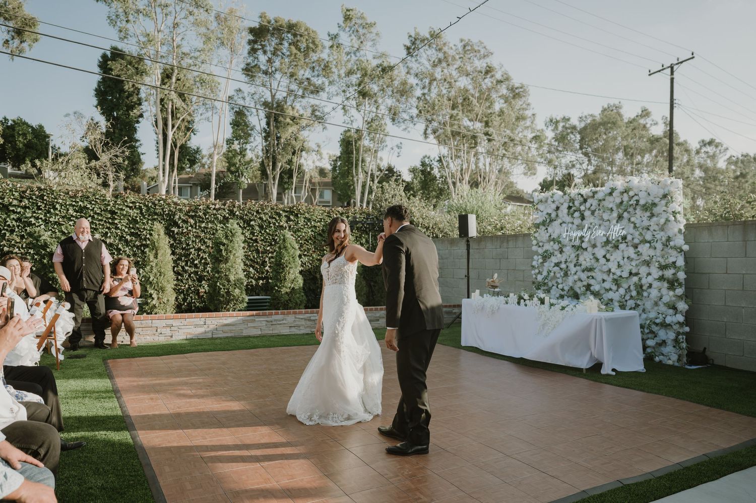 Bride and groom share their first dance outdoors on a wooden floor surrounded by lush greenery at a wedding reception.