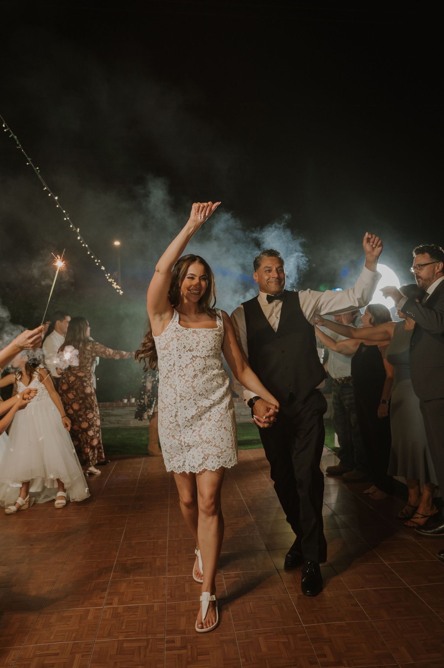 Bride and groom exit wedding reception through sparkler tunnel at night, with guests cheering around them.