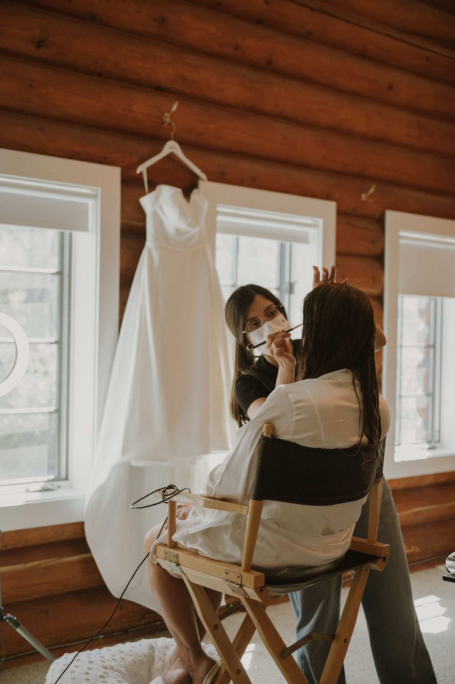 Bride getting makeup done in a log cabin with wedding dress hanging in background.