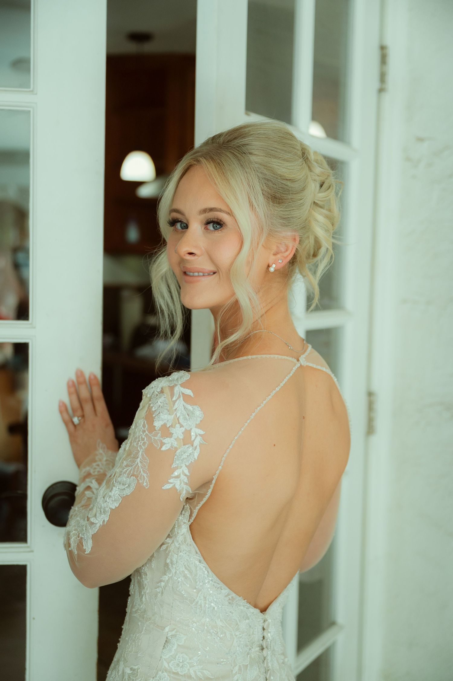 Bride in backless lace wedding gown with updo hairstyle, smiling over shoulder by white French doors.