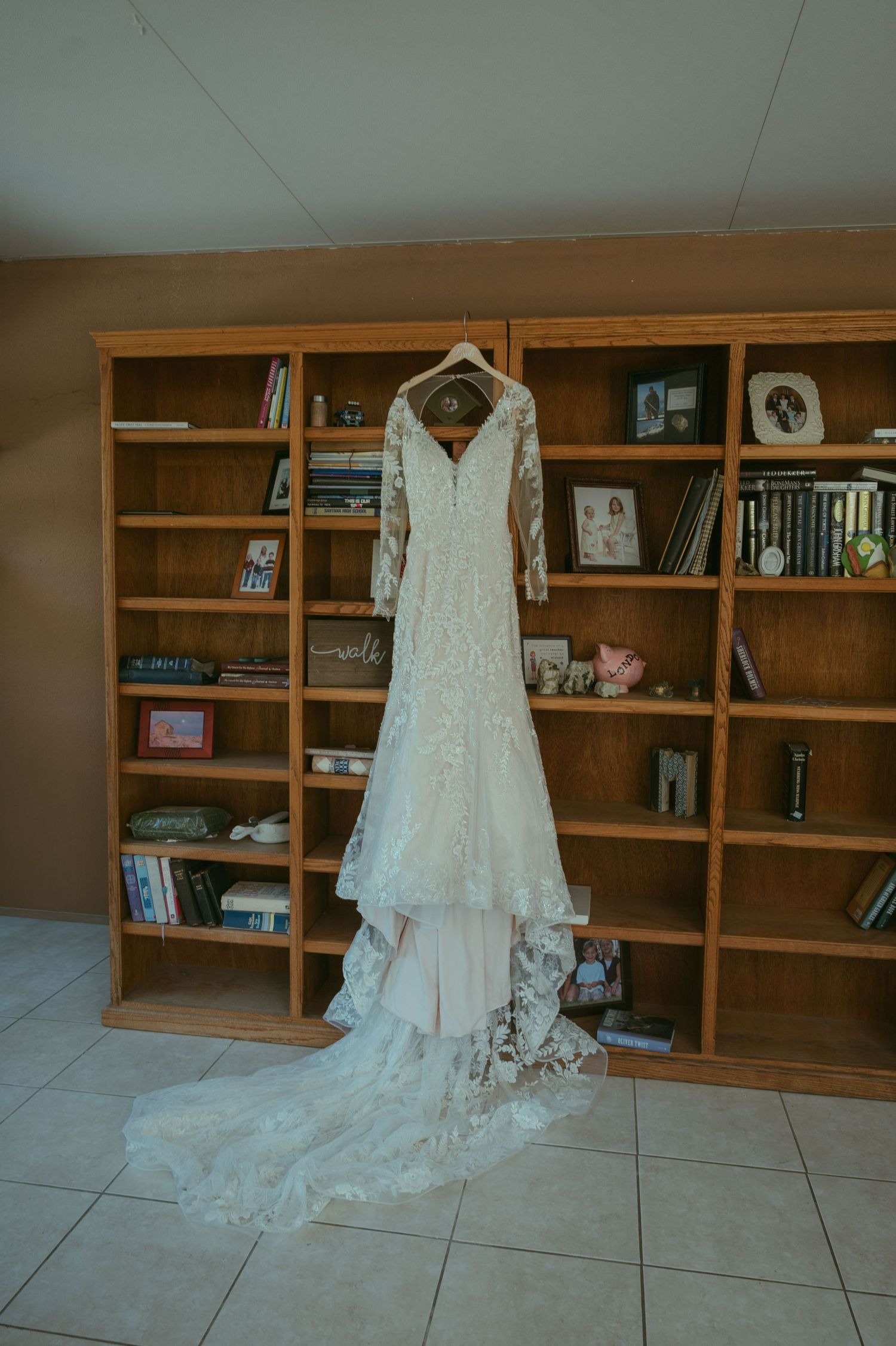 Elegant lace wedding dress with long sleeves and train hanging on wooden bookshelves in a home library.