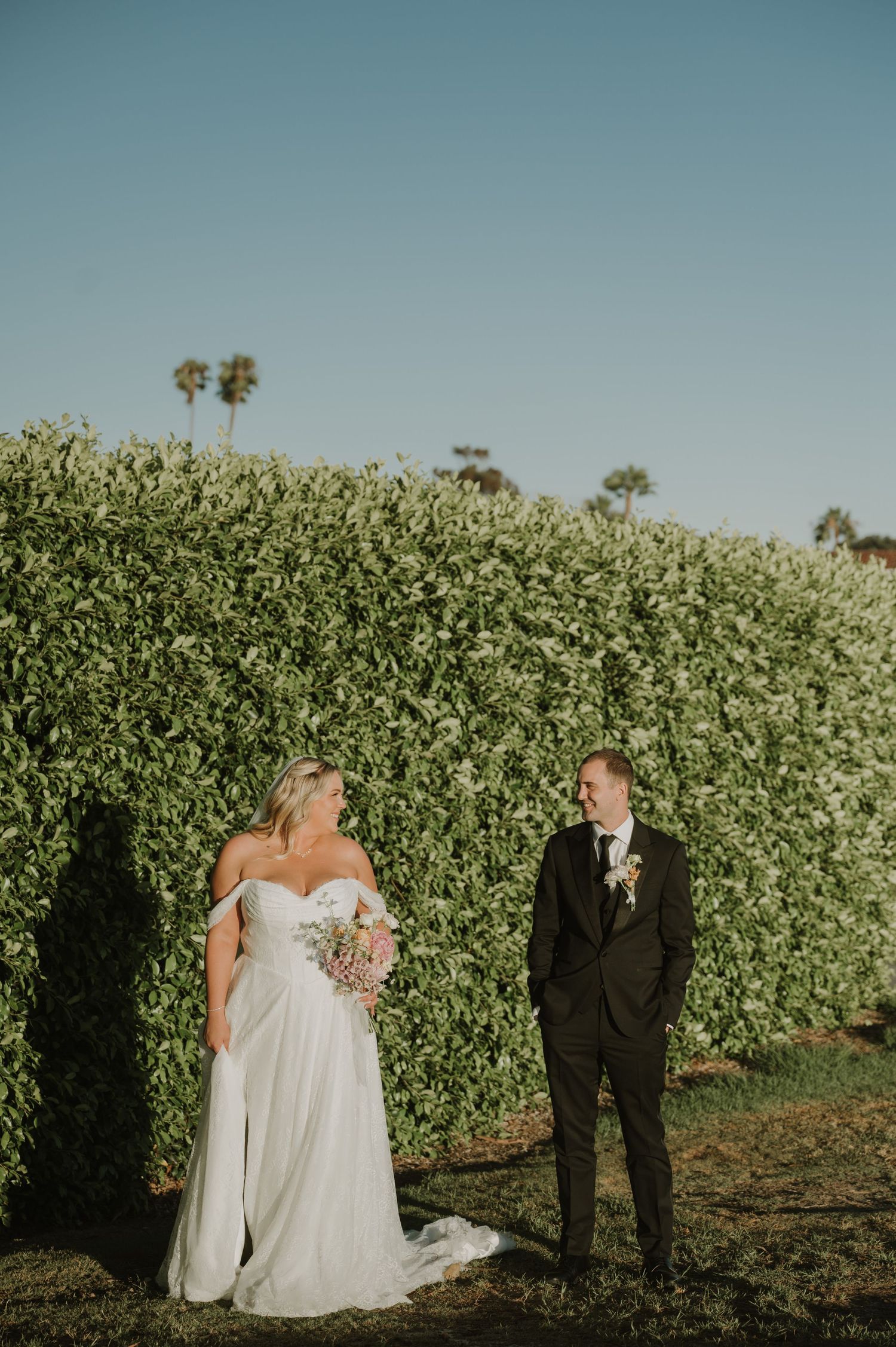 Bride in white off-shoulder gown and groom in black tuxedo stand before a lush green hedge on their wedding day.