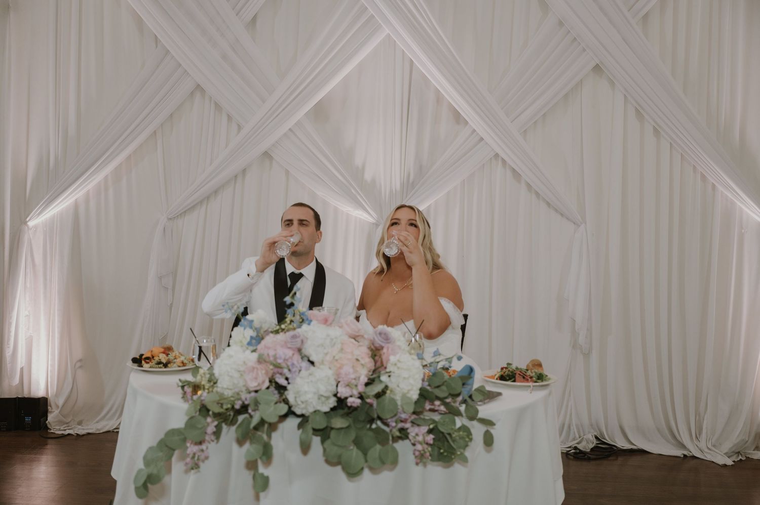 Bride and groom toast at sweetheart table adorned with lush floral centerpiece.