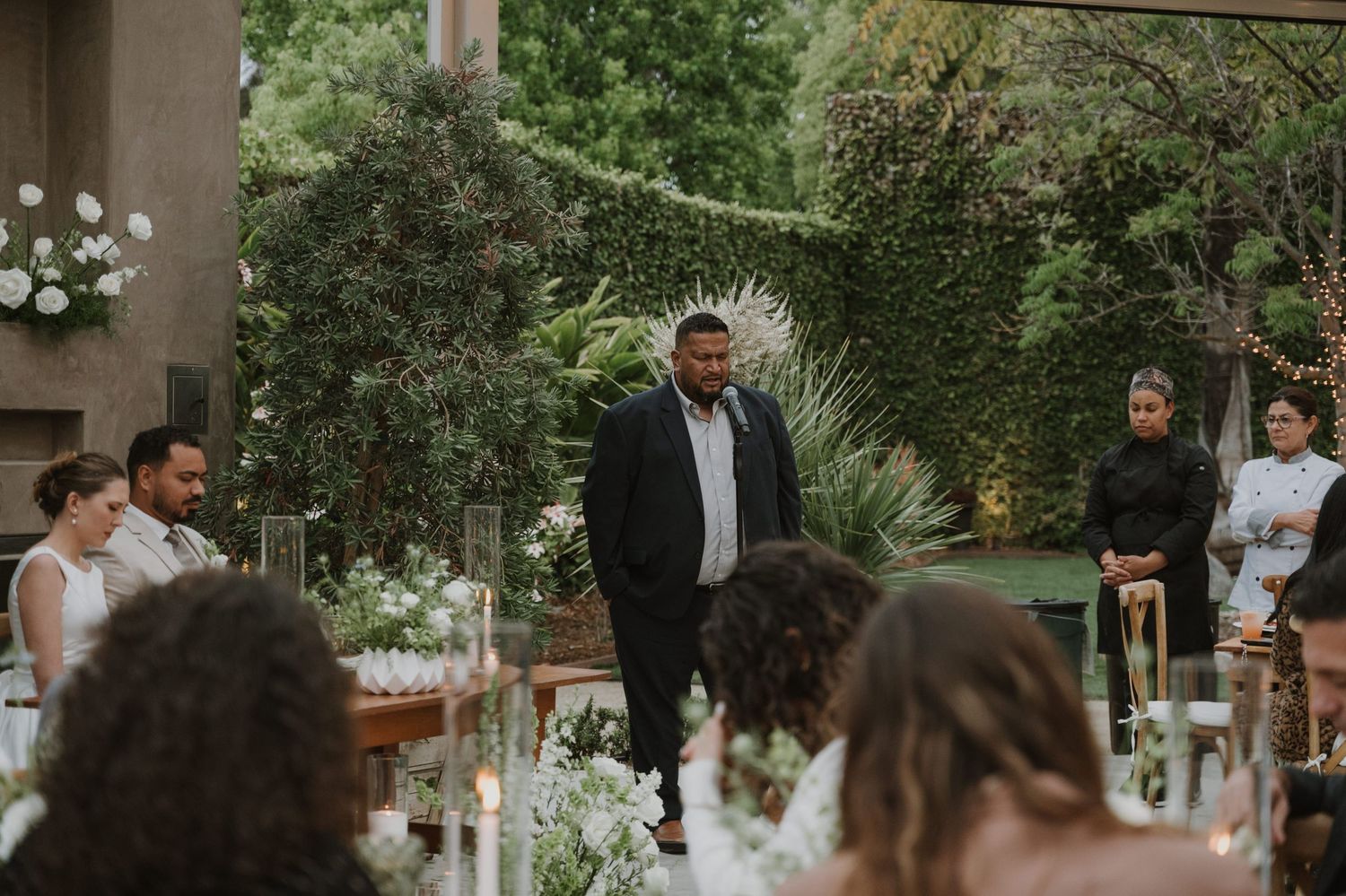 Man in black suit walking down outdoor wedding aisle surrounded by lush greenery and candlelit tables.