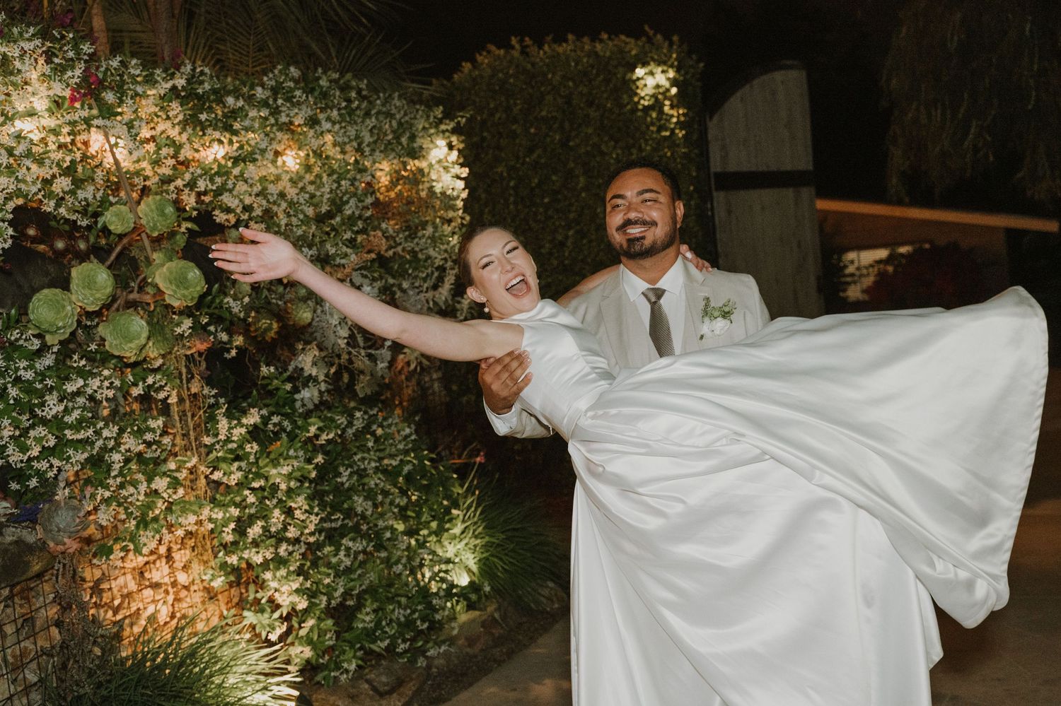 Groom lifting smiling bride in white gown in front of lush green floral wall at night.