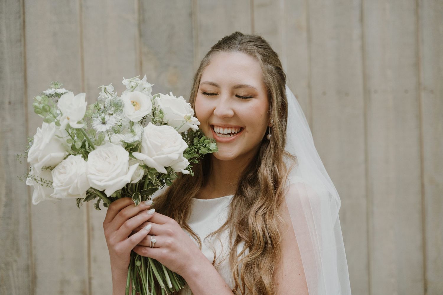 Smiling bride in white veil holding elegant white rose bouquet, laughing with eyes closed on her wedding day.