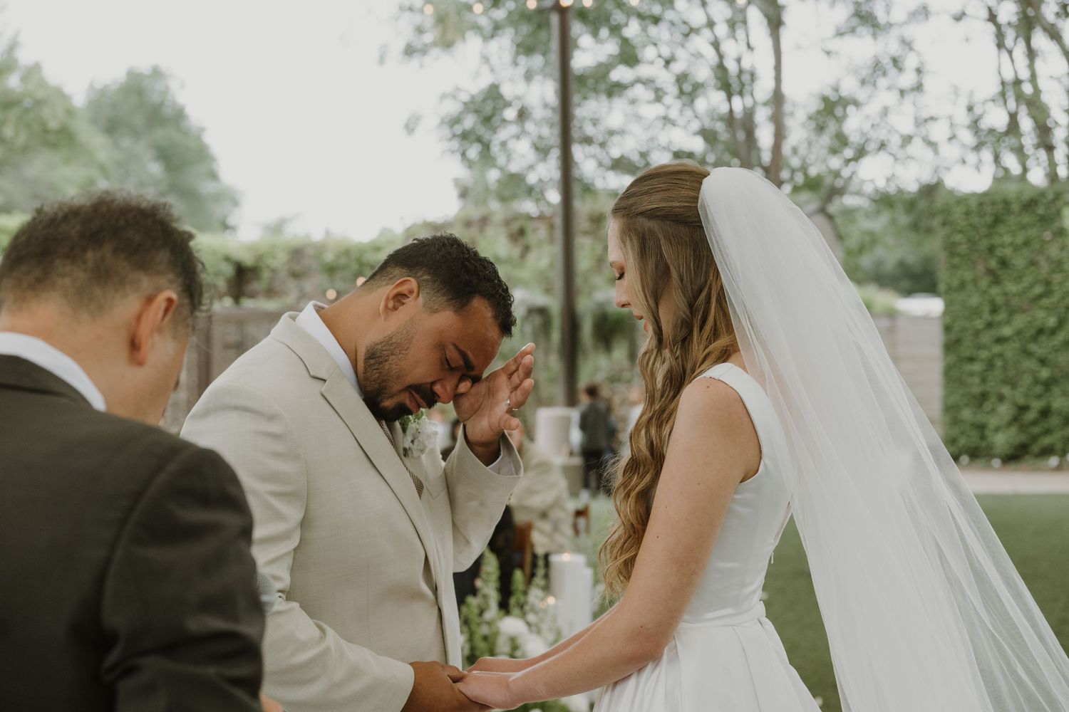 Emotional groom wiping tears during outdoor wedding ceremony while bride holds hands with officiant.