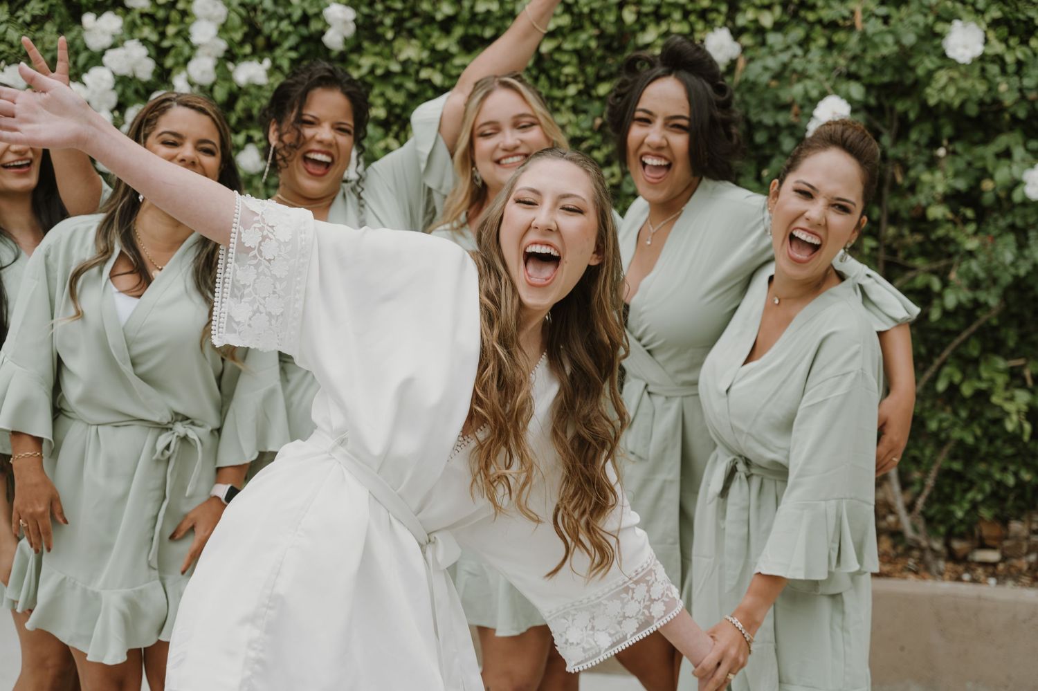 Bride in white robe laughing with bridesmaids in sage green robes during wedding preparations outdoors.