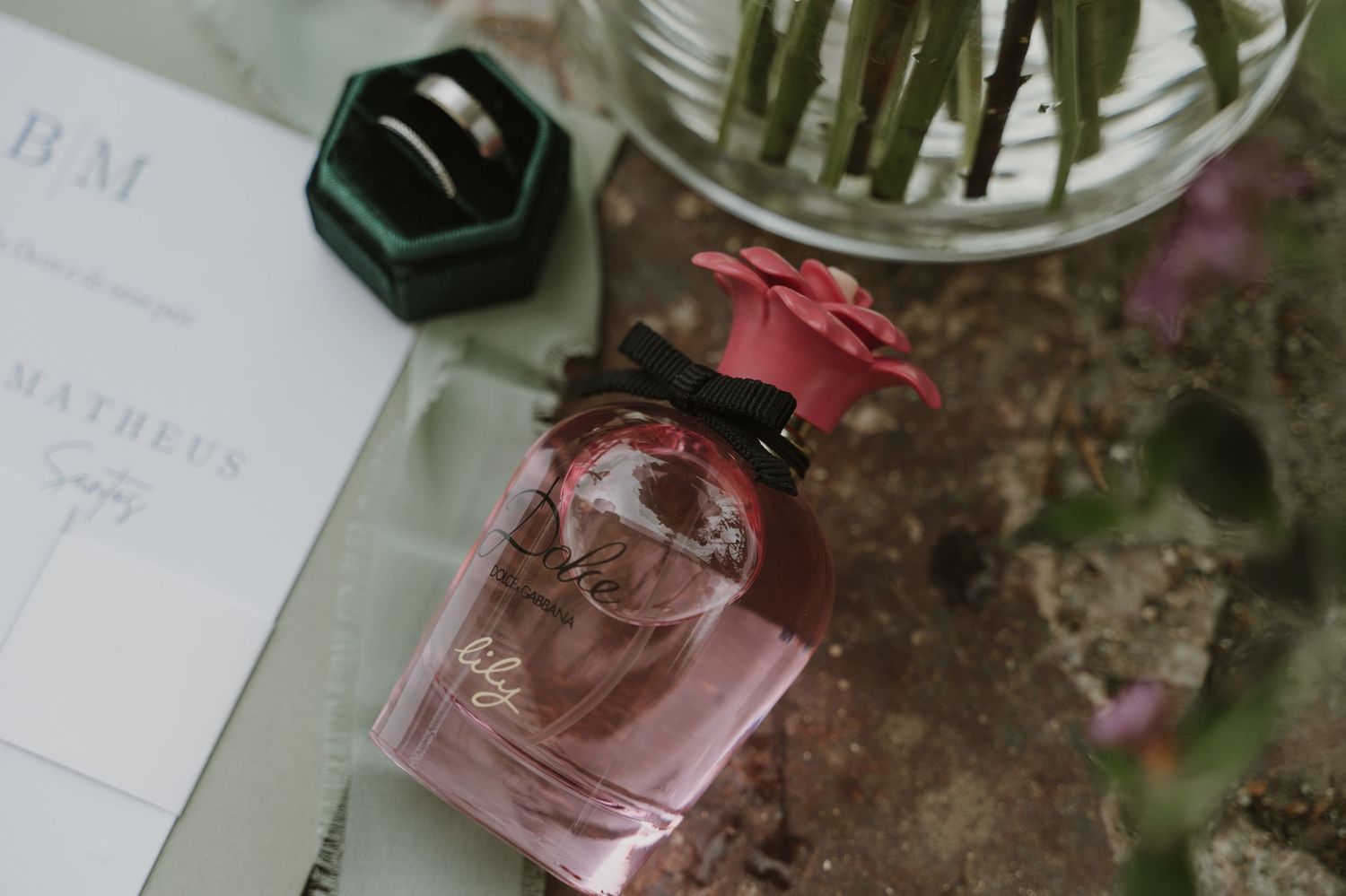 Flat lay of pink floral perfume bottle beside wedding stationery and green velvet ring box.