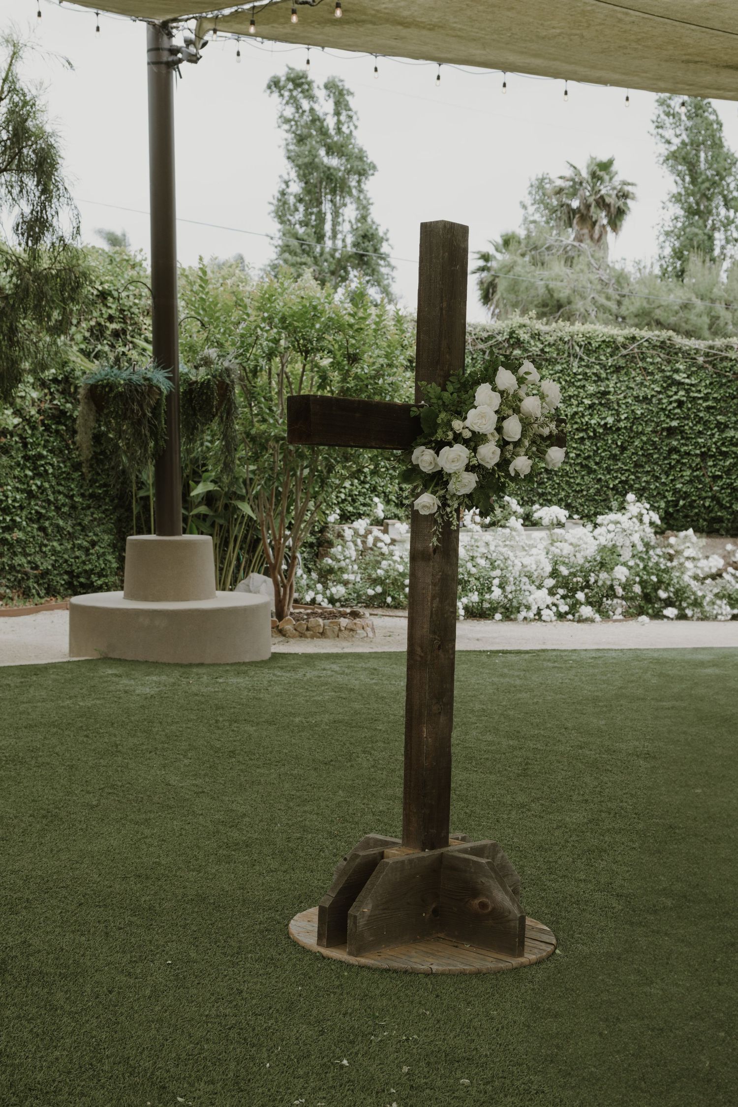Rustic wooden cross decorated with white roses on green lawn at an outdoor wedding ceremony venue.