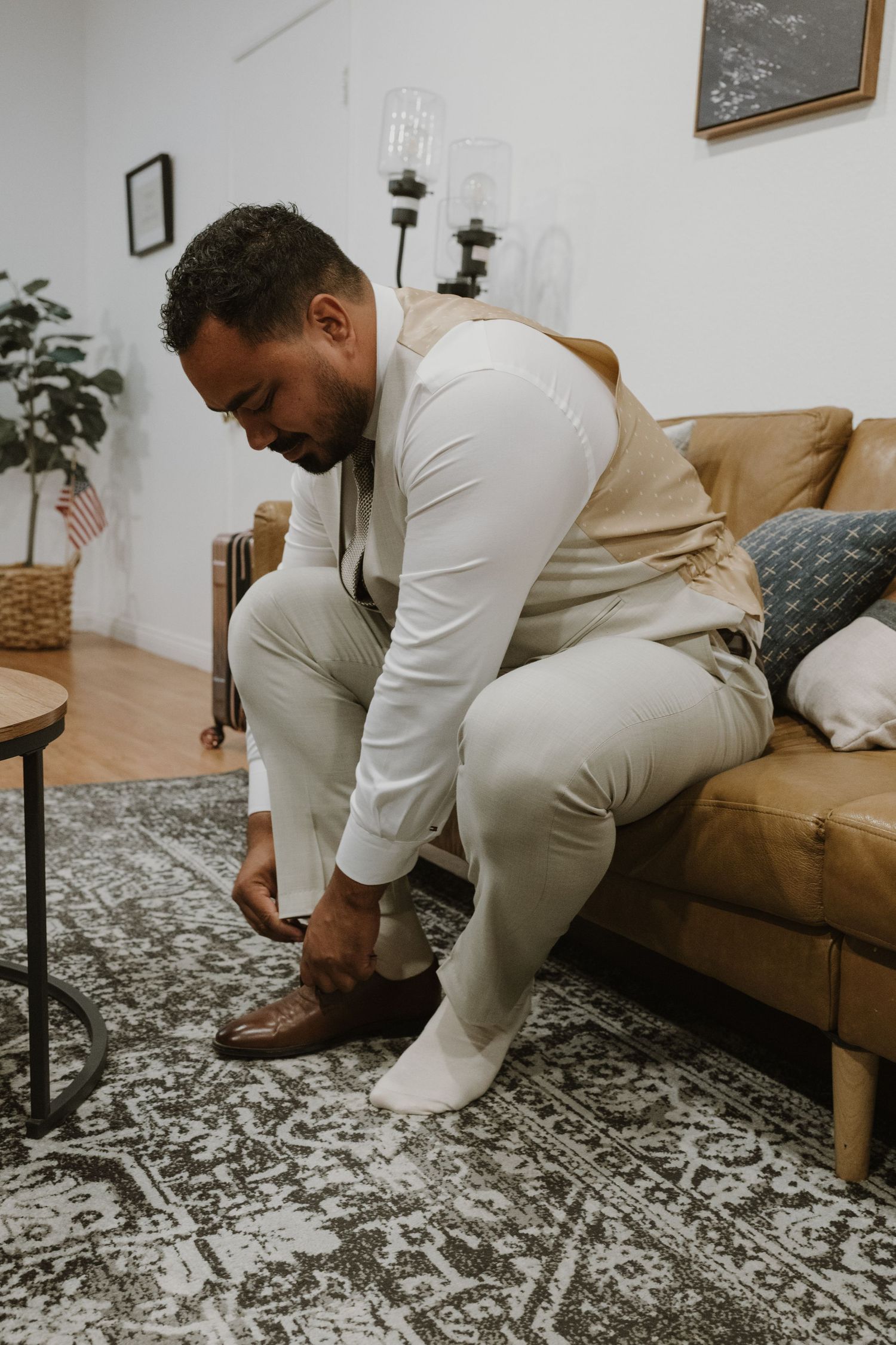 A groom in a white shirt and tan vest sits on a leather sofa, putting on brown dress shoes before his wedding.