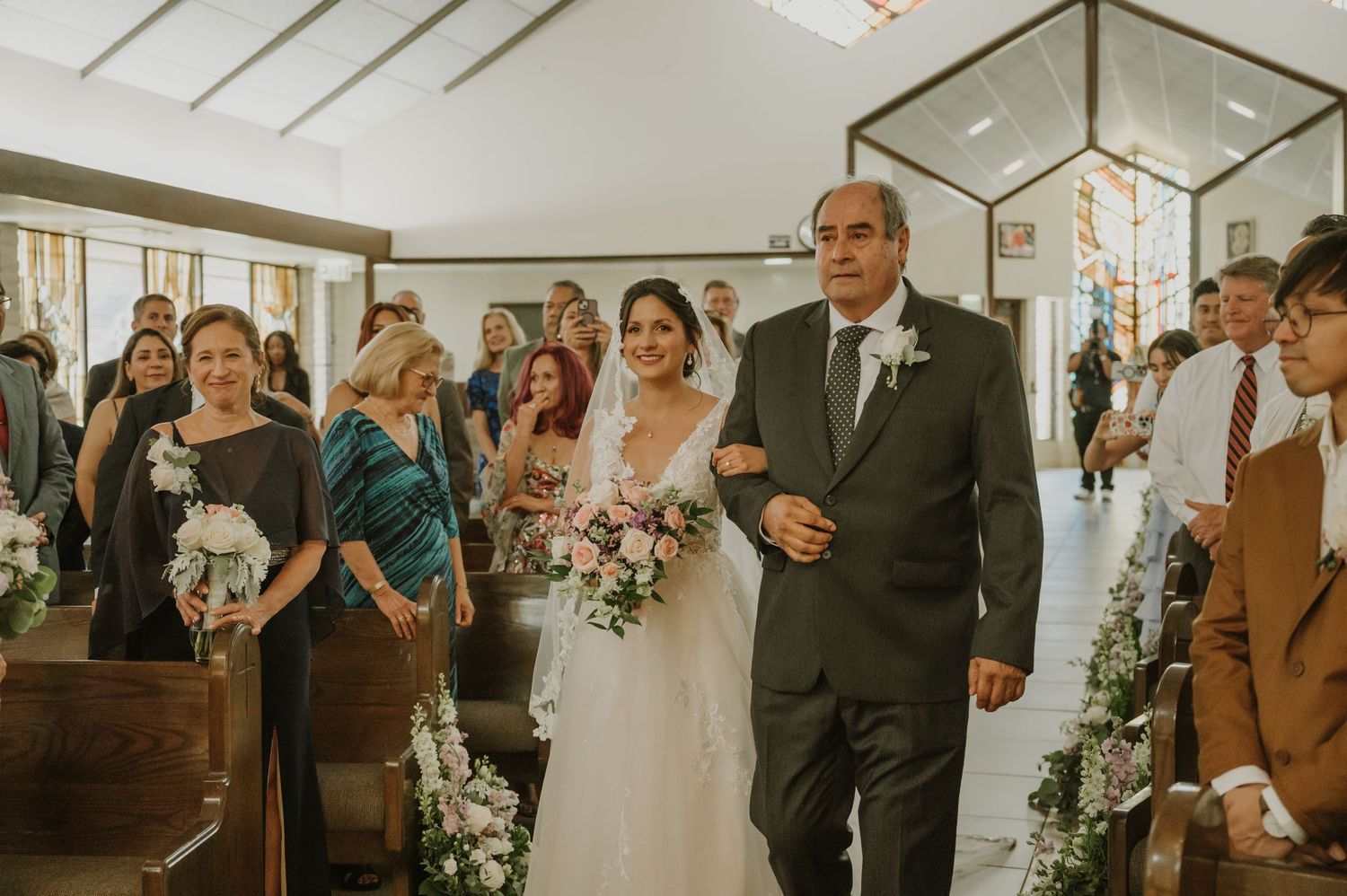Bride in white gown walks down church aisle escorted by older man, guests watching and holding bouquets.