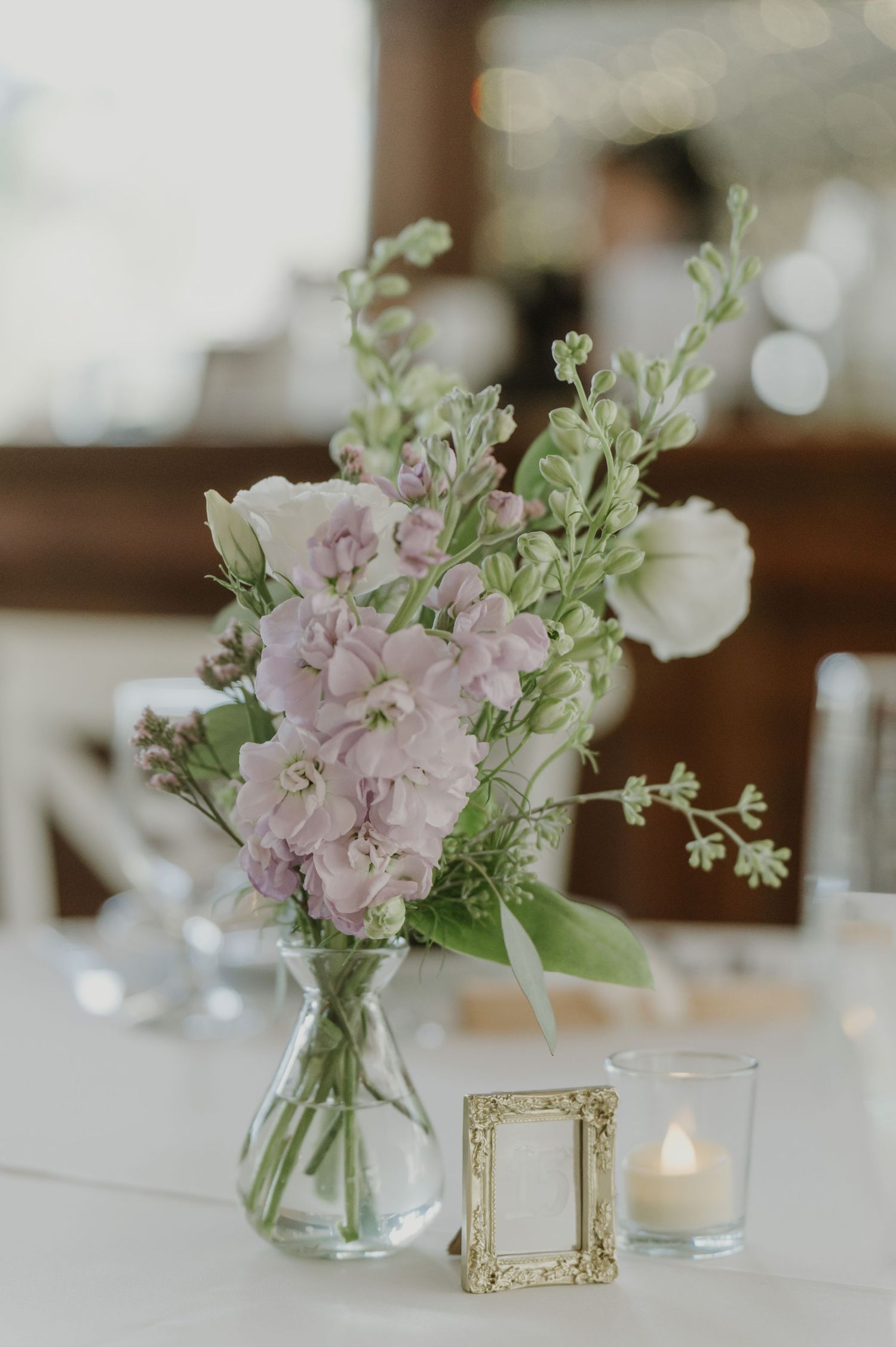 Glass vase with pink and white flowers beside a gold frame and candle on a white wedding reception table.