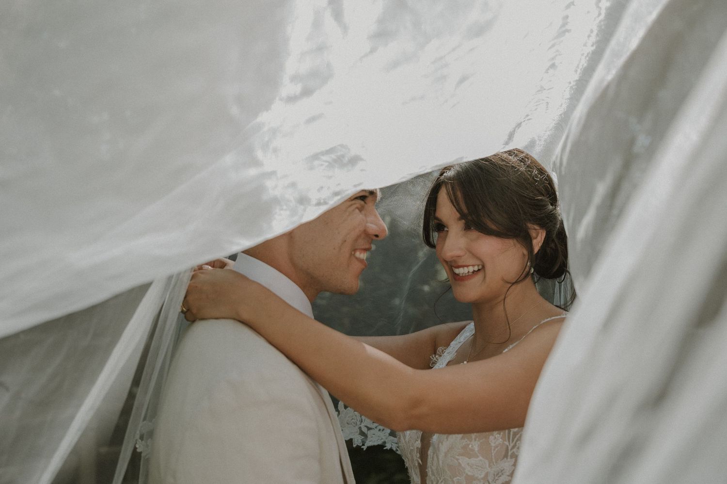 A bride and groom share a romantic moment under a flowing white wedding veil, smiling at each other outdoors.