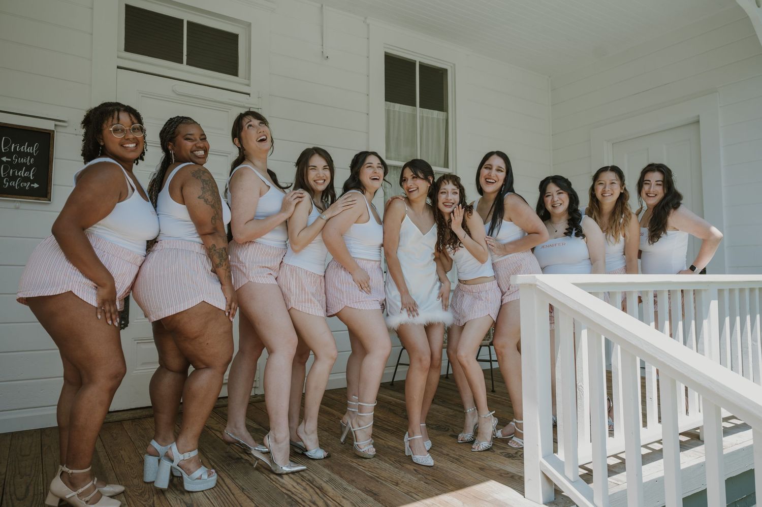 Group of bridesmaids in white matching outfits posing and laughing on a wooden porch railing.