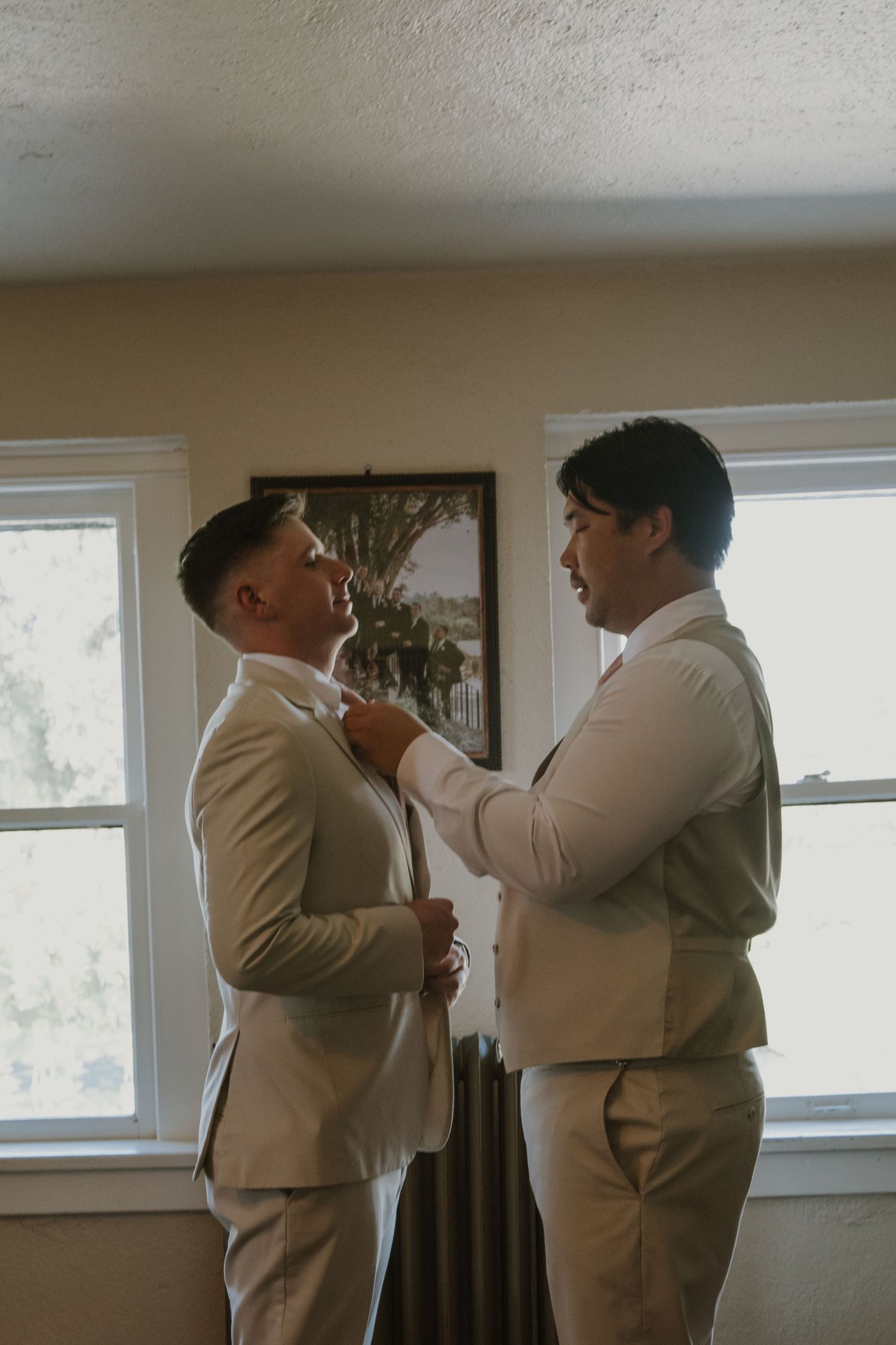 Two grooms in matching cream suits help each other with ties while getting ready near bright windows on wedding day.