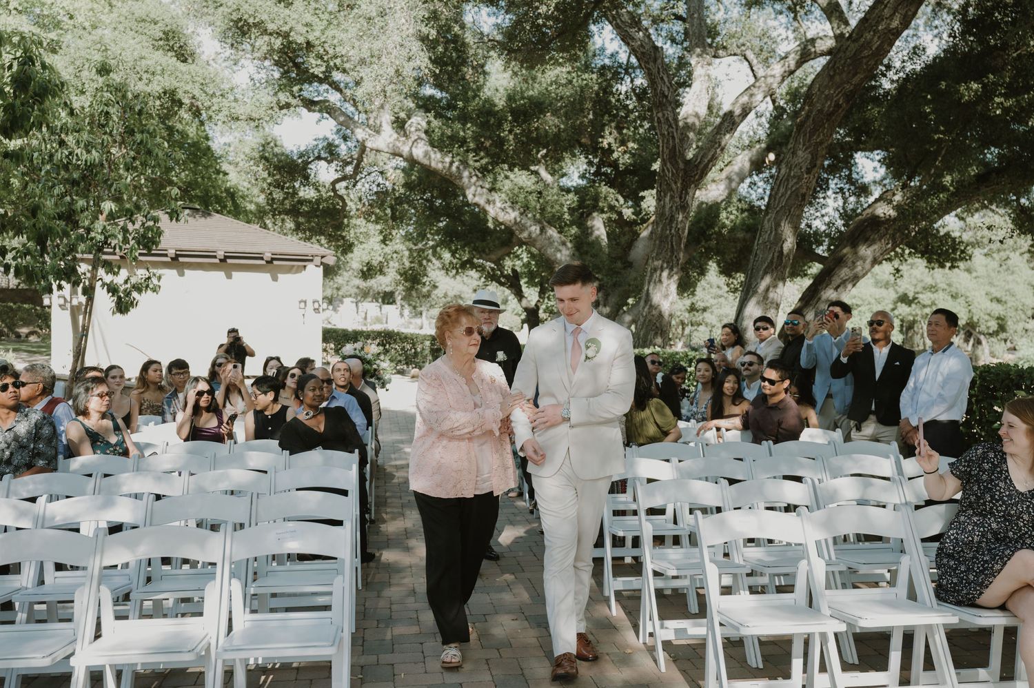 Groom walks down outdoor wedding aisle with mother, surrounded by seated guests under large oak trees.