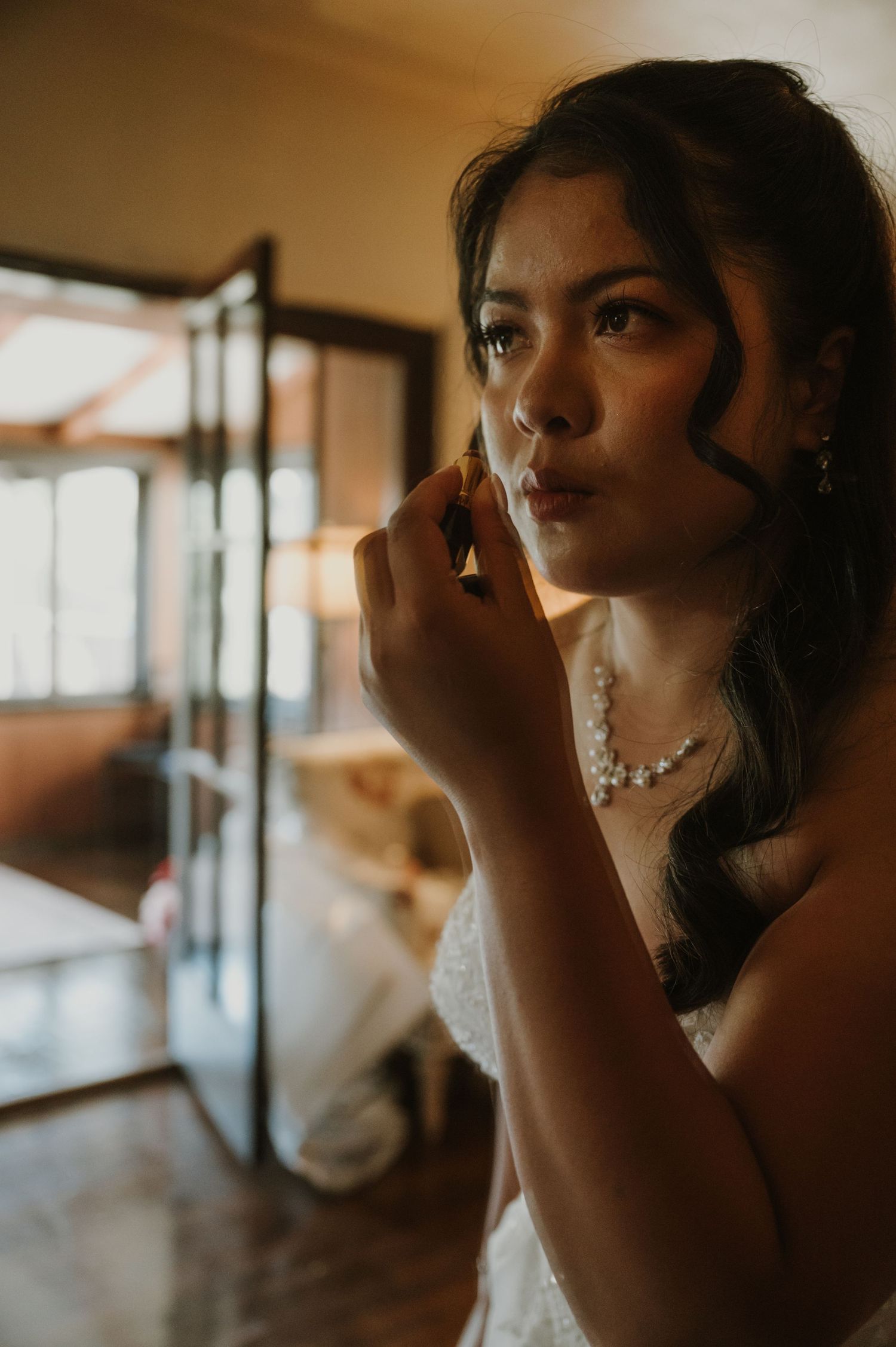 Bride in white dress applying makeup, wearing pearl necklace and earrings, preparing for her wedding day.
