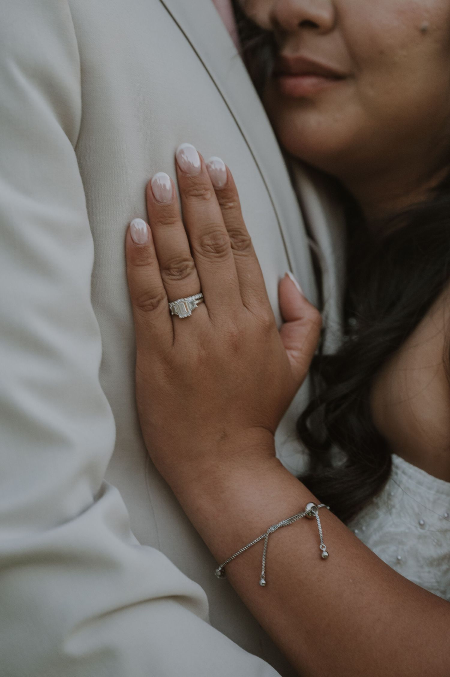 Close-up of brides diamond wedding ring and bracelet as she embraces groom in white suit.