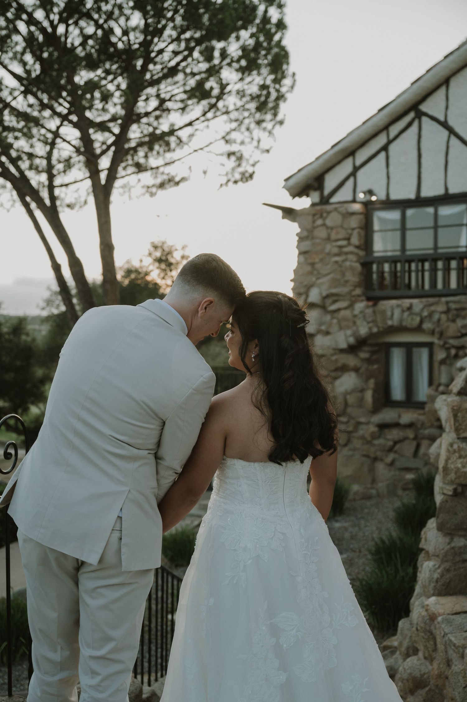 Bride and groom share a tender moment walking toward a rustic stone venue at sunset.