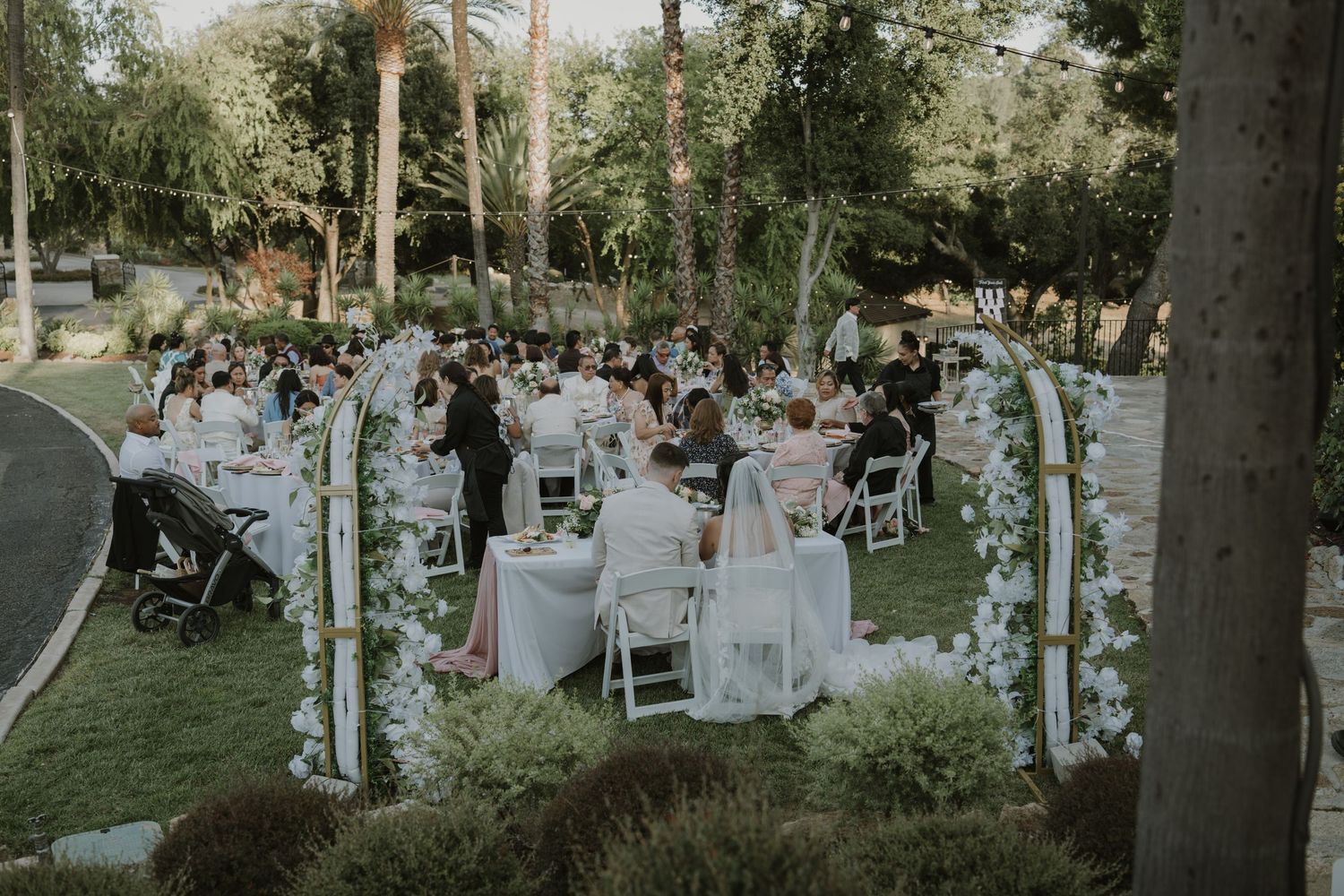 Elegant outdoor wedding reception with white chairs and floral arches on lush green lawn surrounded by tall trees.