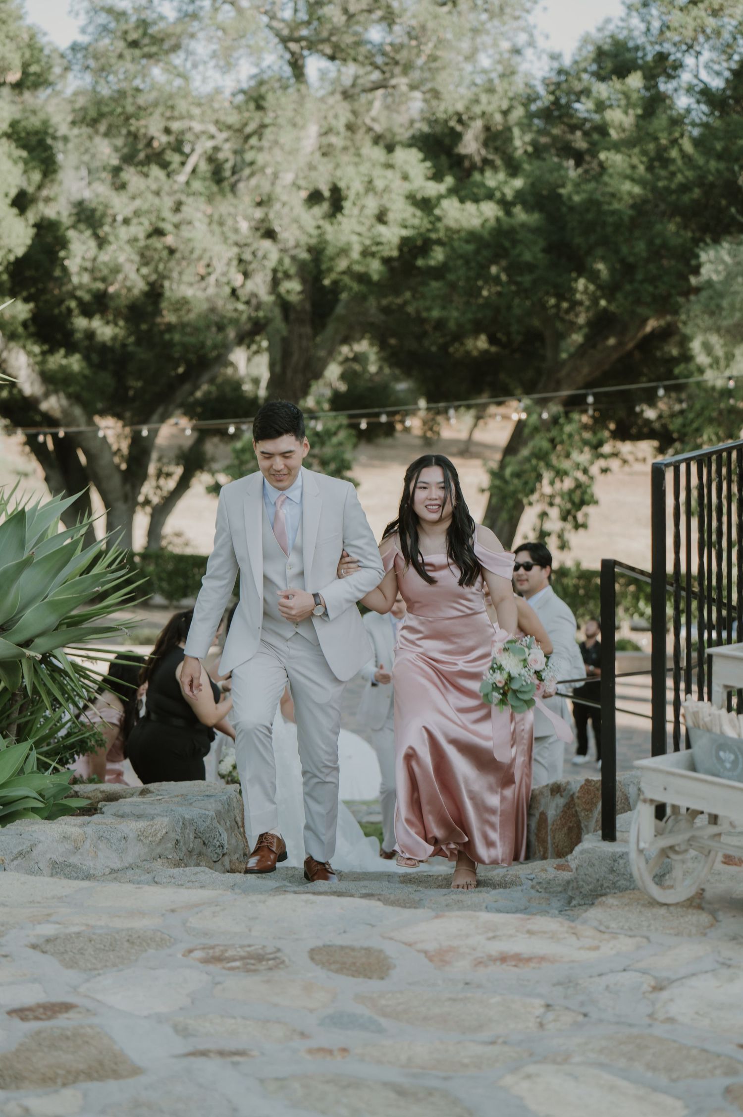 Smiling couple in formal attire walking up stone steps at an outdoor wedding venue surrounded by trees.