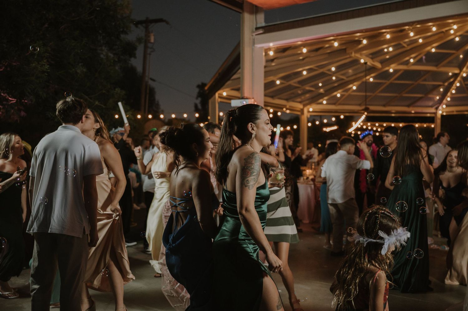 Wedding guests dance under string lights at lively outdoor evening reception celebration.
