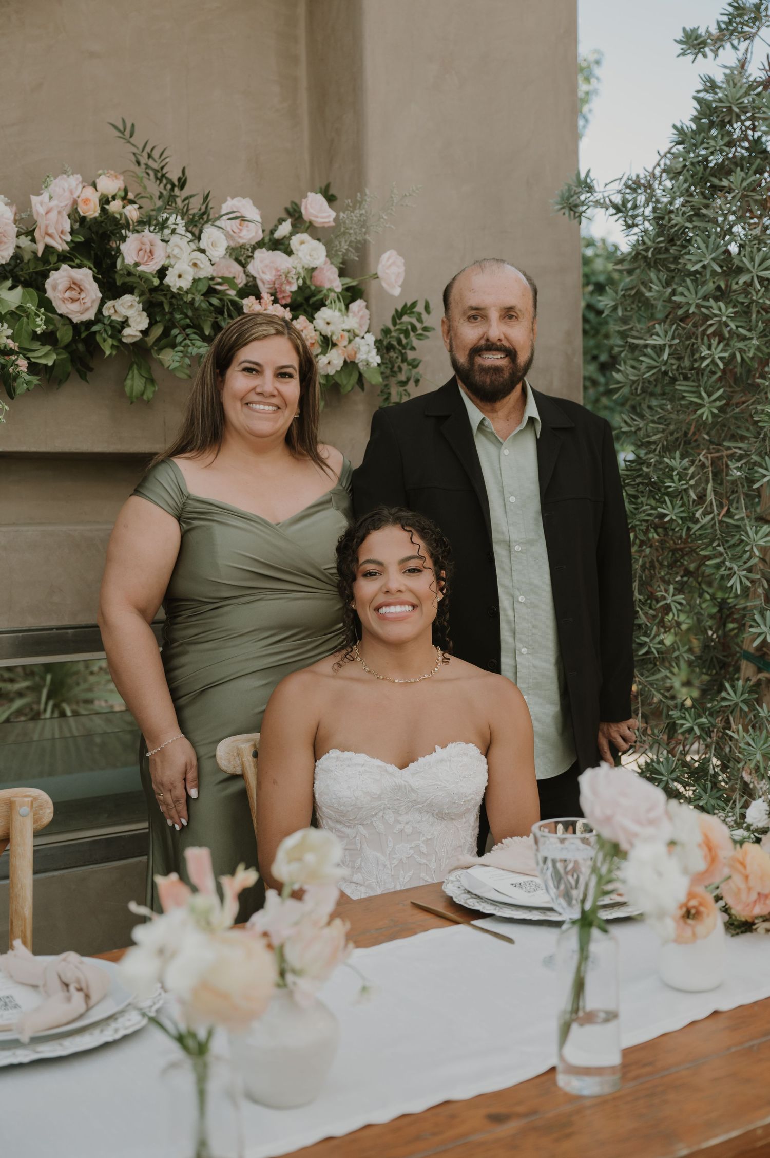 Bride in white strapless gown seated at reception table with parents posing behind floral arrangements.
