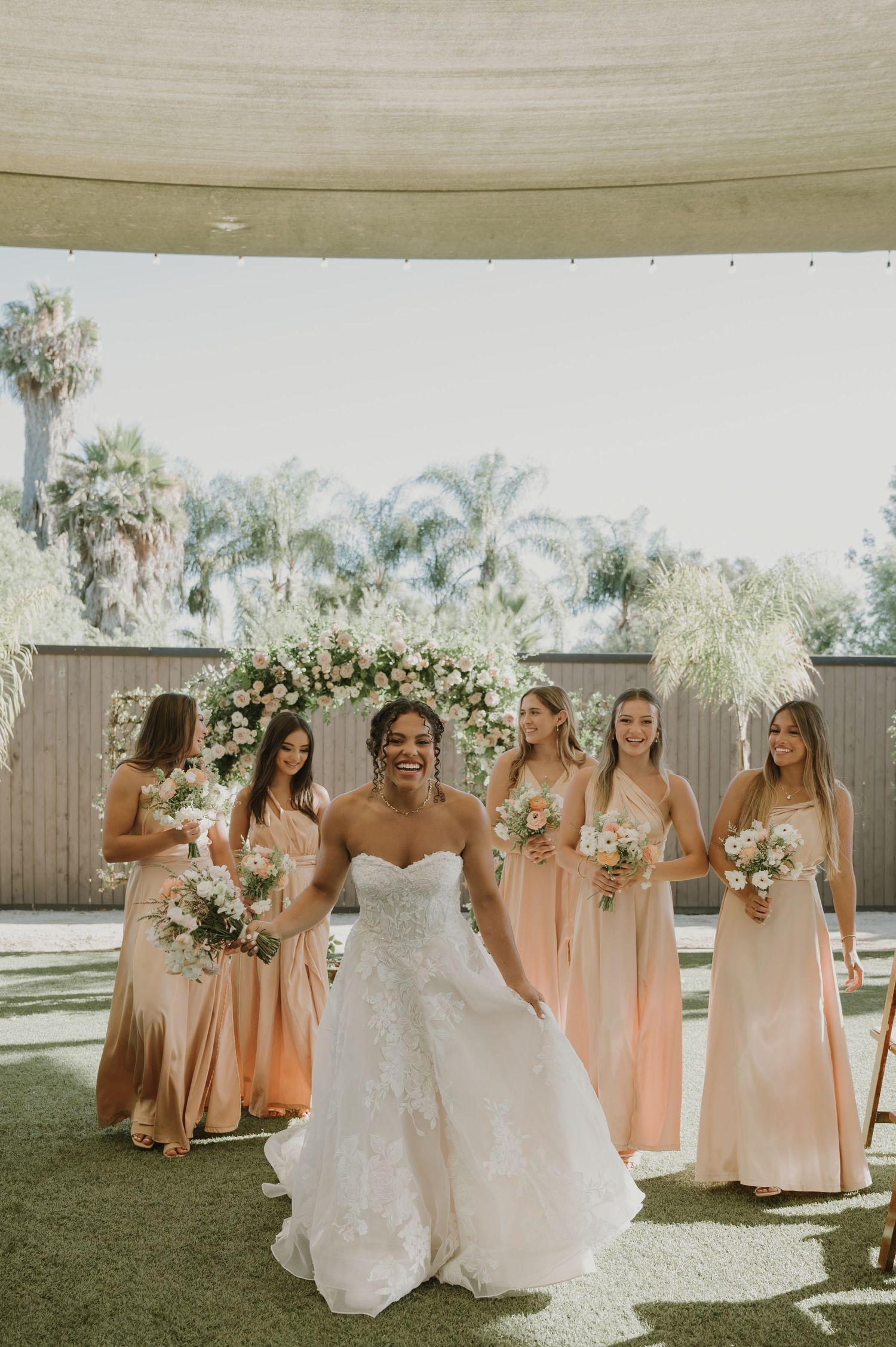 Bride in white lace gown smiles with four bridesmaids in blush dresses holding bouquets outdoors.